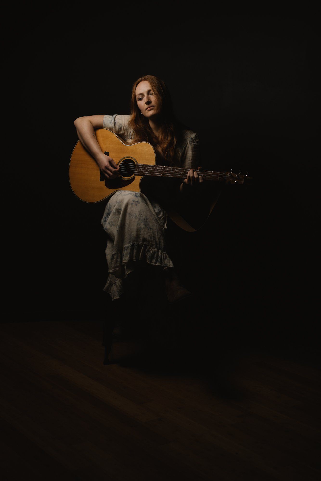Woman with long reddish-brown hair holds an acoustic guitar against a dark background, gazing thoughtfully upward in moody studio lighting