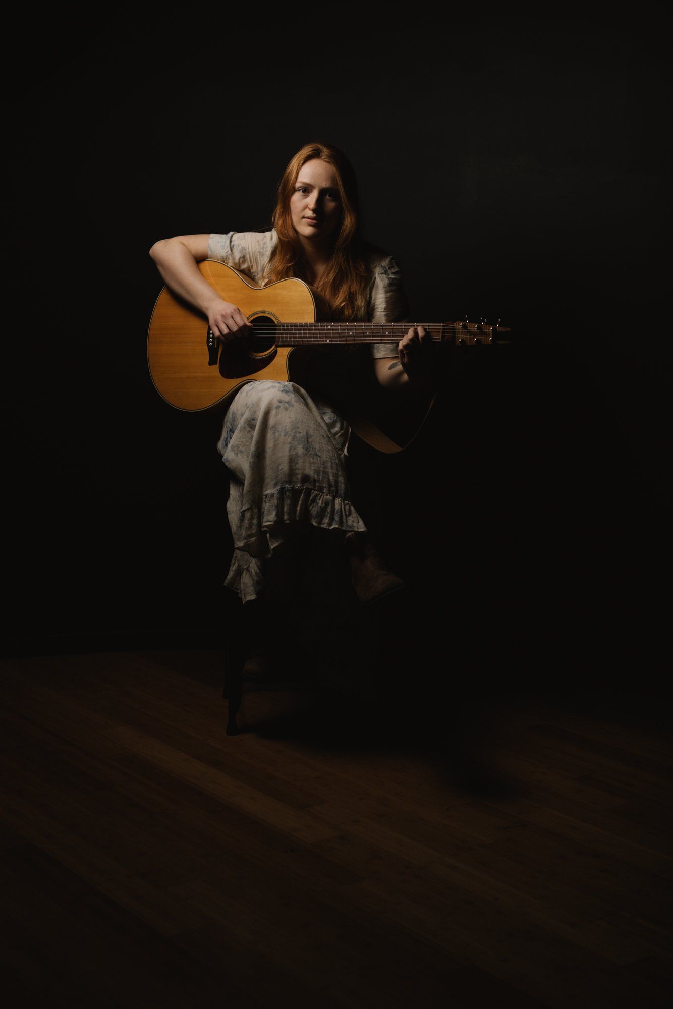 Woman with long reddish-brown hair plays acoustic guitar against a dark background, warmly lit from the side