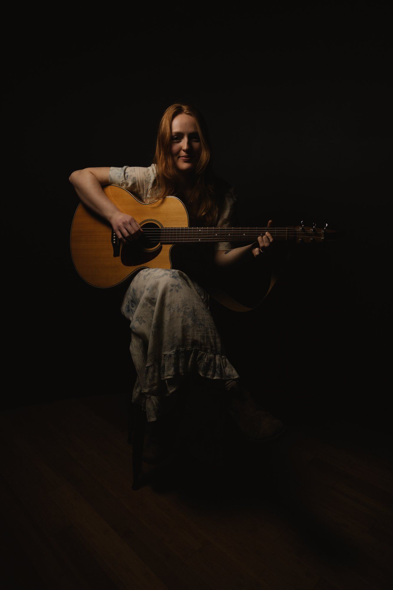 Woman with long reddish-brown hair playing acoustic guitar against a dark background, softly lit