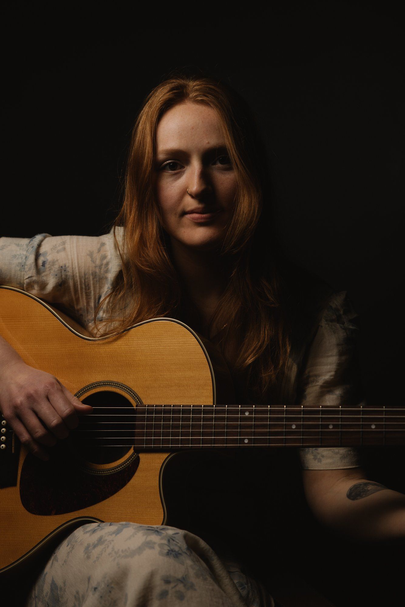 Woman with long reddish-brown hair holding an acoustic guitar against a dark background, warm studio lighting