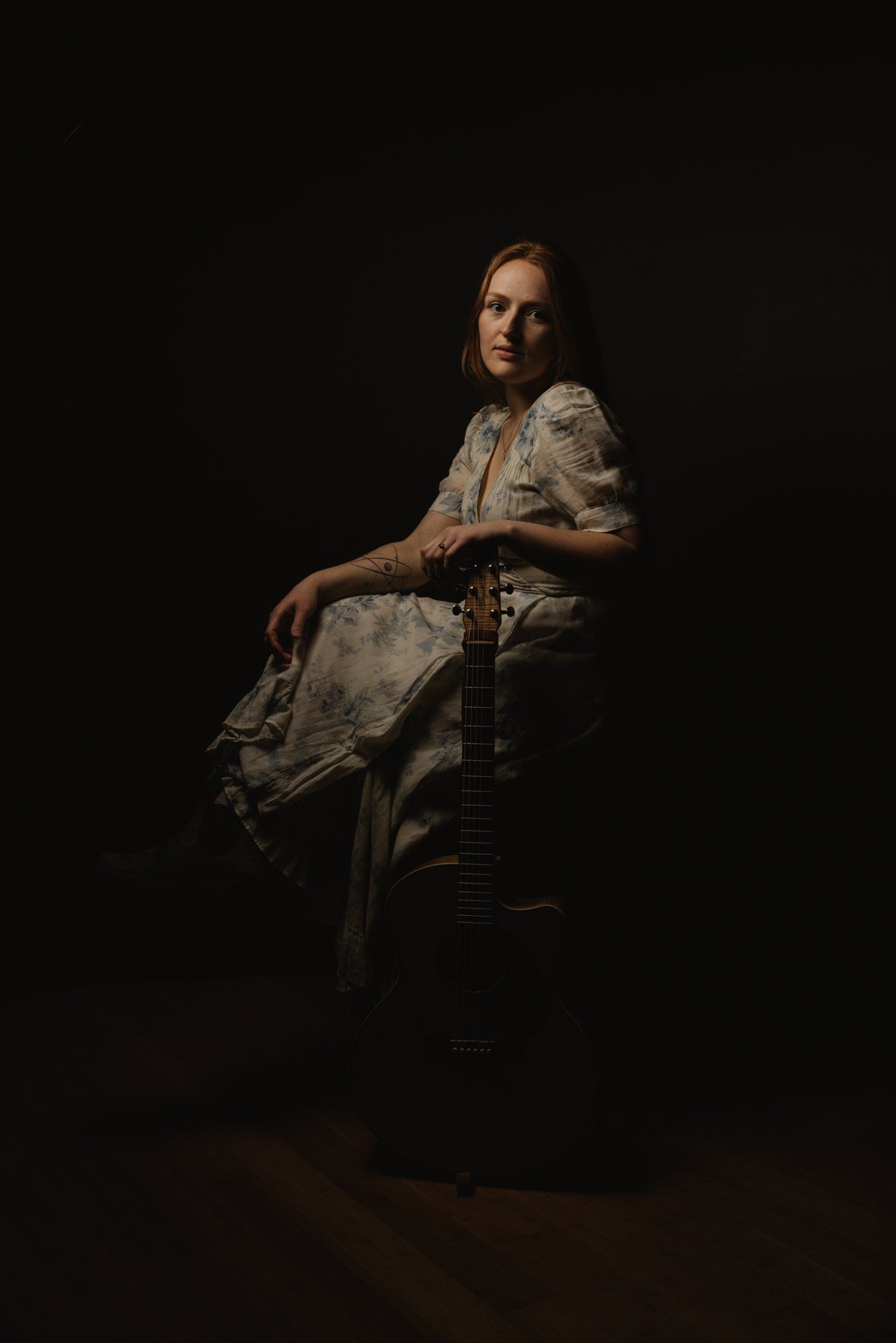 Woman in cream lace dress seated on wooden chair against black background, contemplative gaze, dramatic side lighting