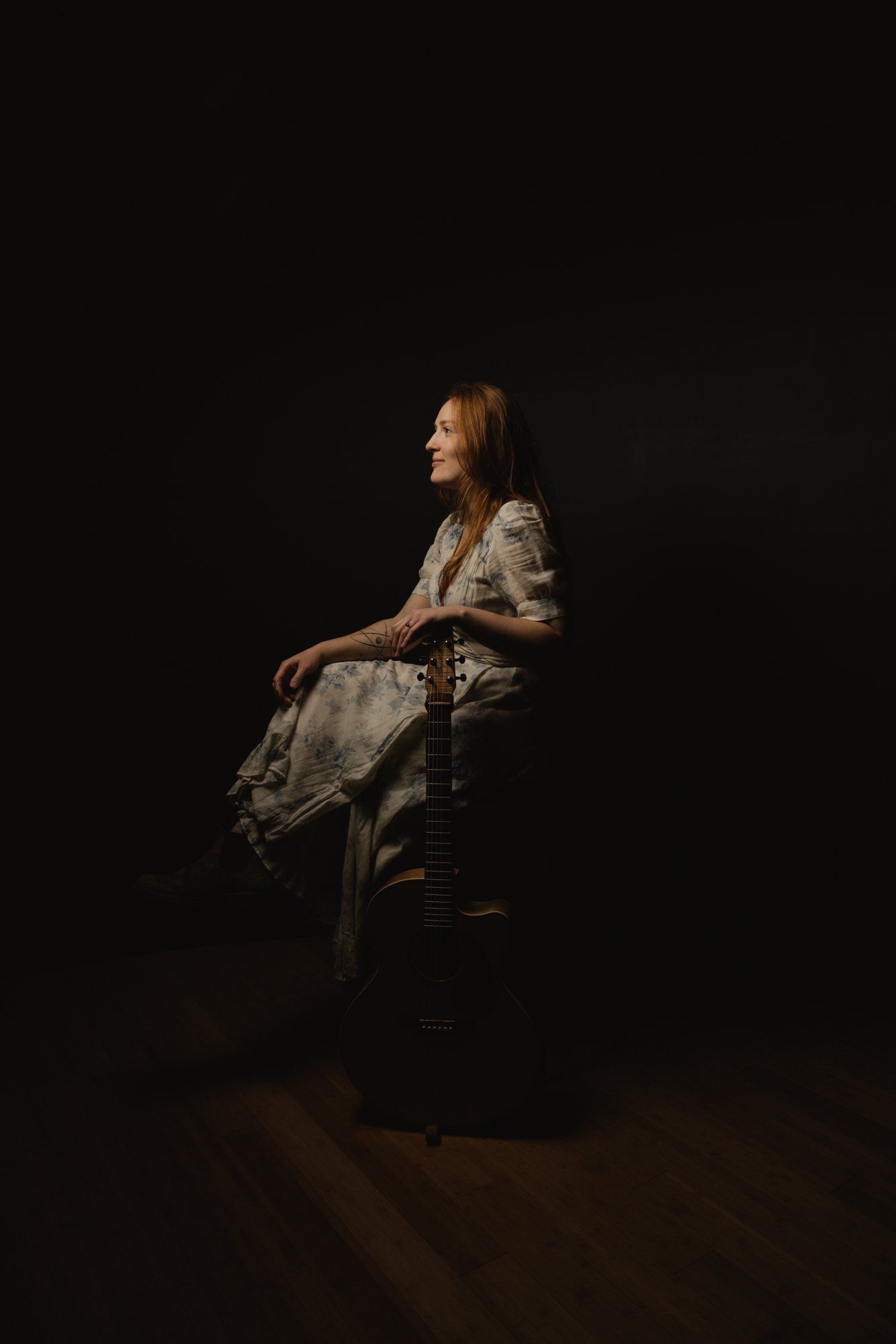 Woman with reddish hair seated on a wooden stool against black background, wearing a linen shirt and flowing skirt, gazing thoughtfully to the side in warm s…
