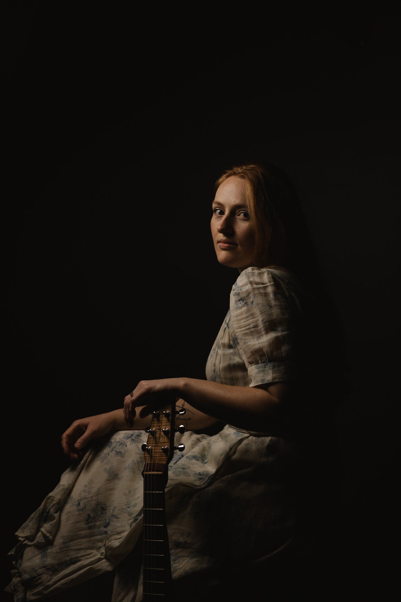 Woman in light linen shirt holding acoustic guitar, seated against black background with dramatic side lighting