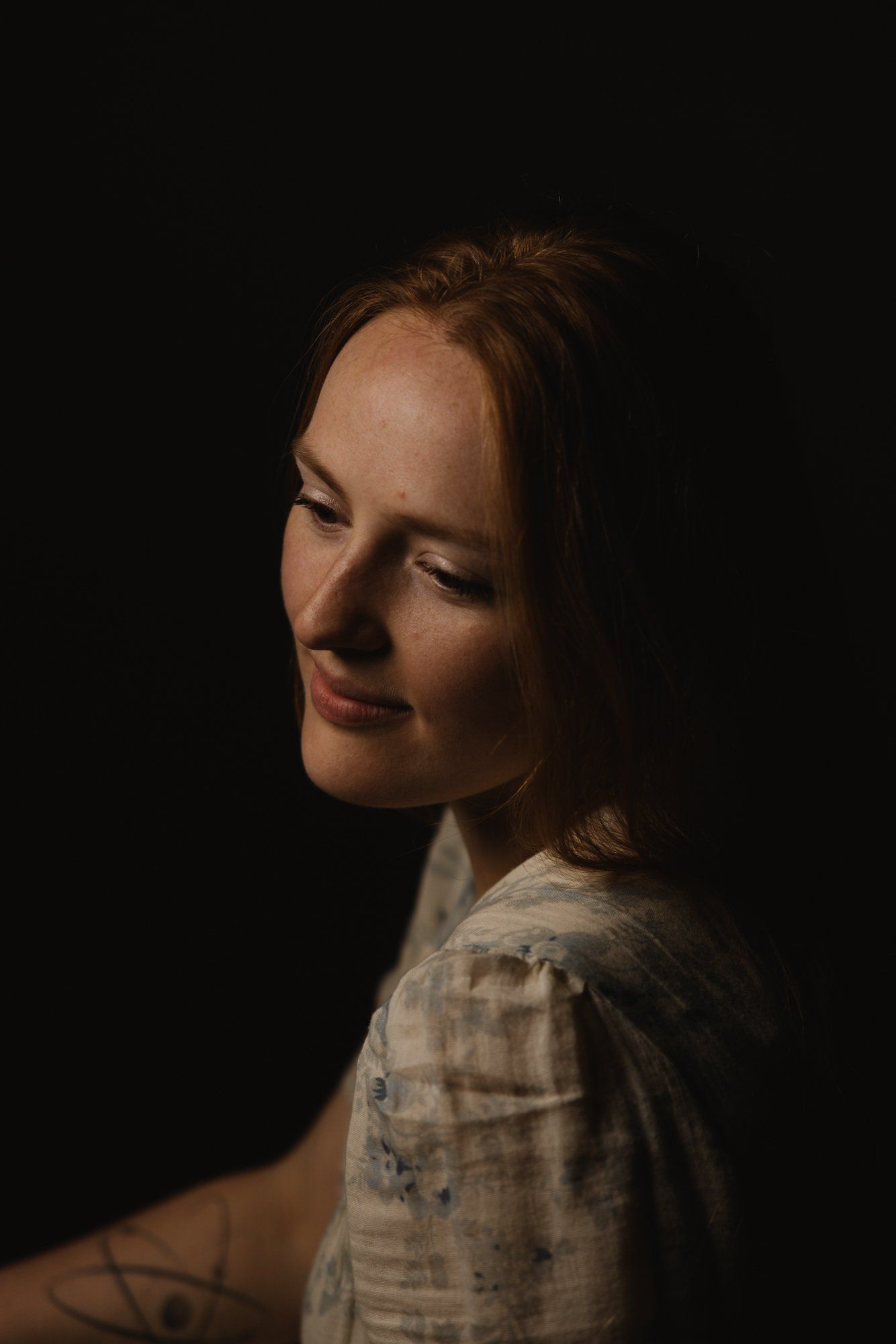 Woman with reddish-brown hair in soft profile against black background, warm dramatic lighting illuminating her face and shoulder