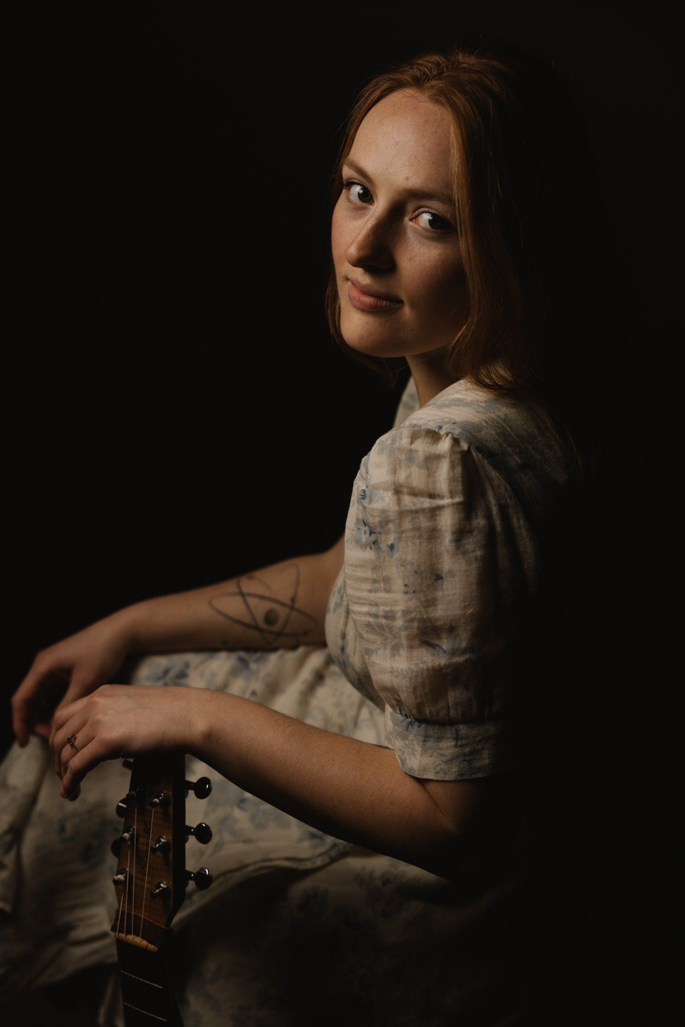 Woman in light plaid shirt holding acoustic guitar, gazing over shoulder against dark background with warm side lighting