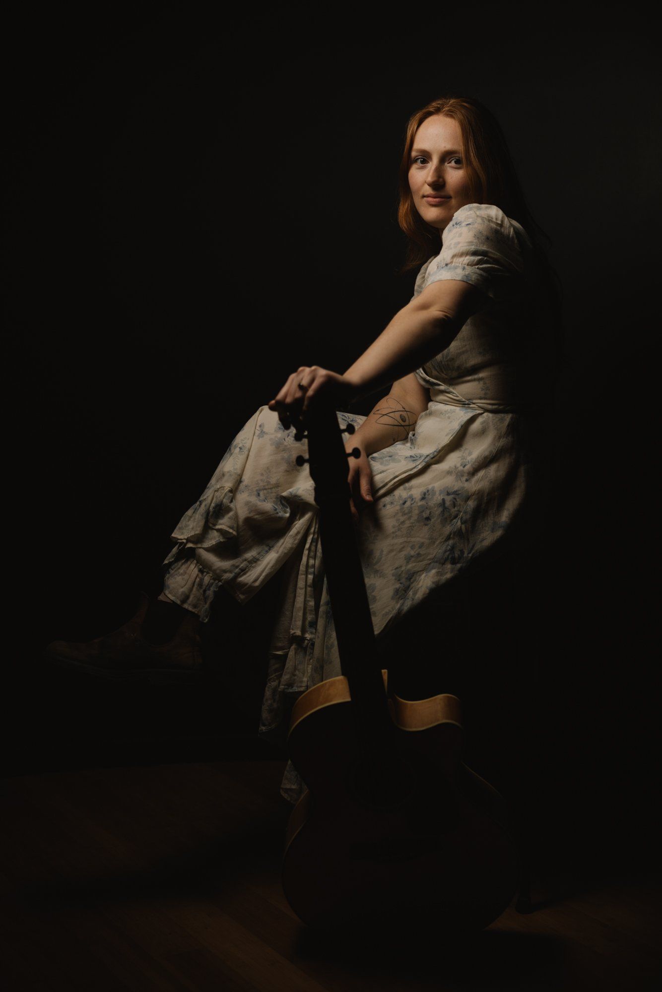 Woman in cream linen shirt and tan pants seated against black background, holding denim jacket, warm side lighting