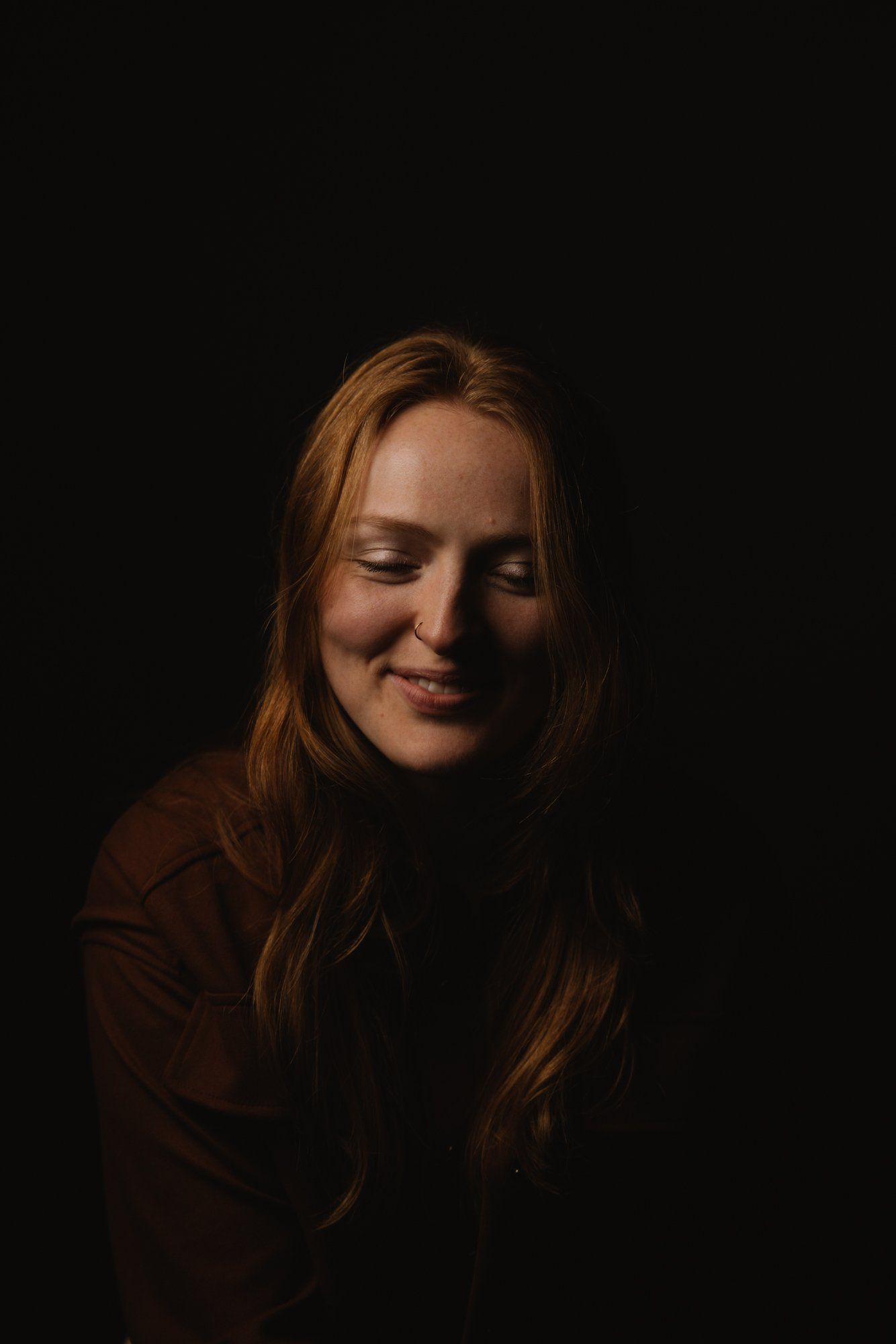Woman with reddish-brown hair smiling softly against dark background, lit from warm side light