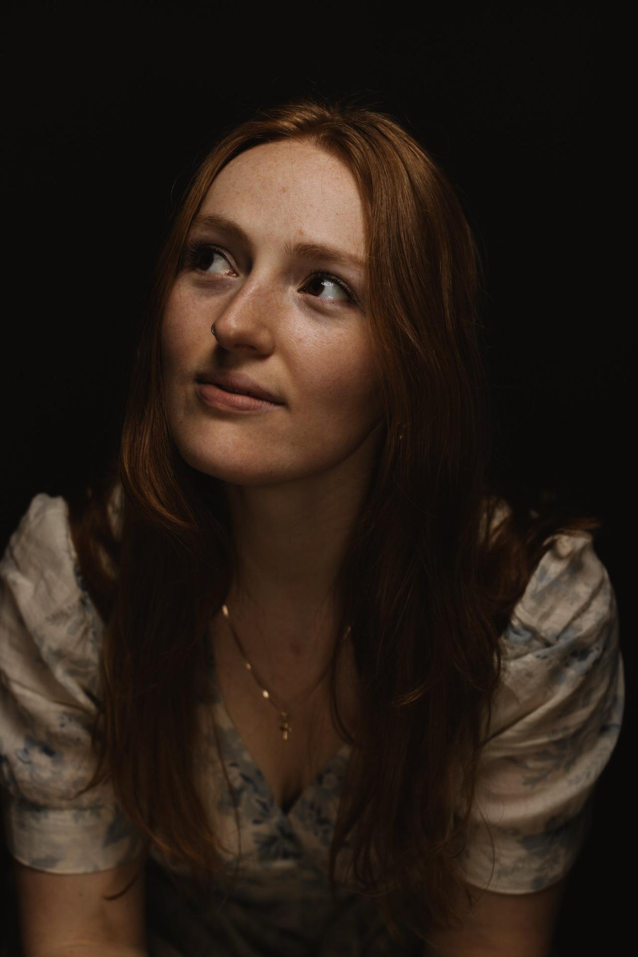 Woman with long reddish-brown hair gazes upward in profile against dark background, sidelit