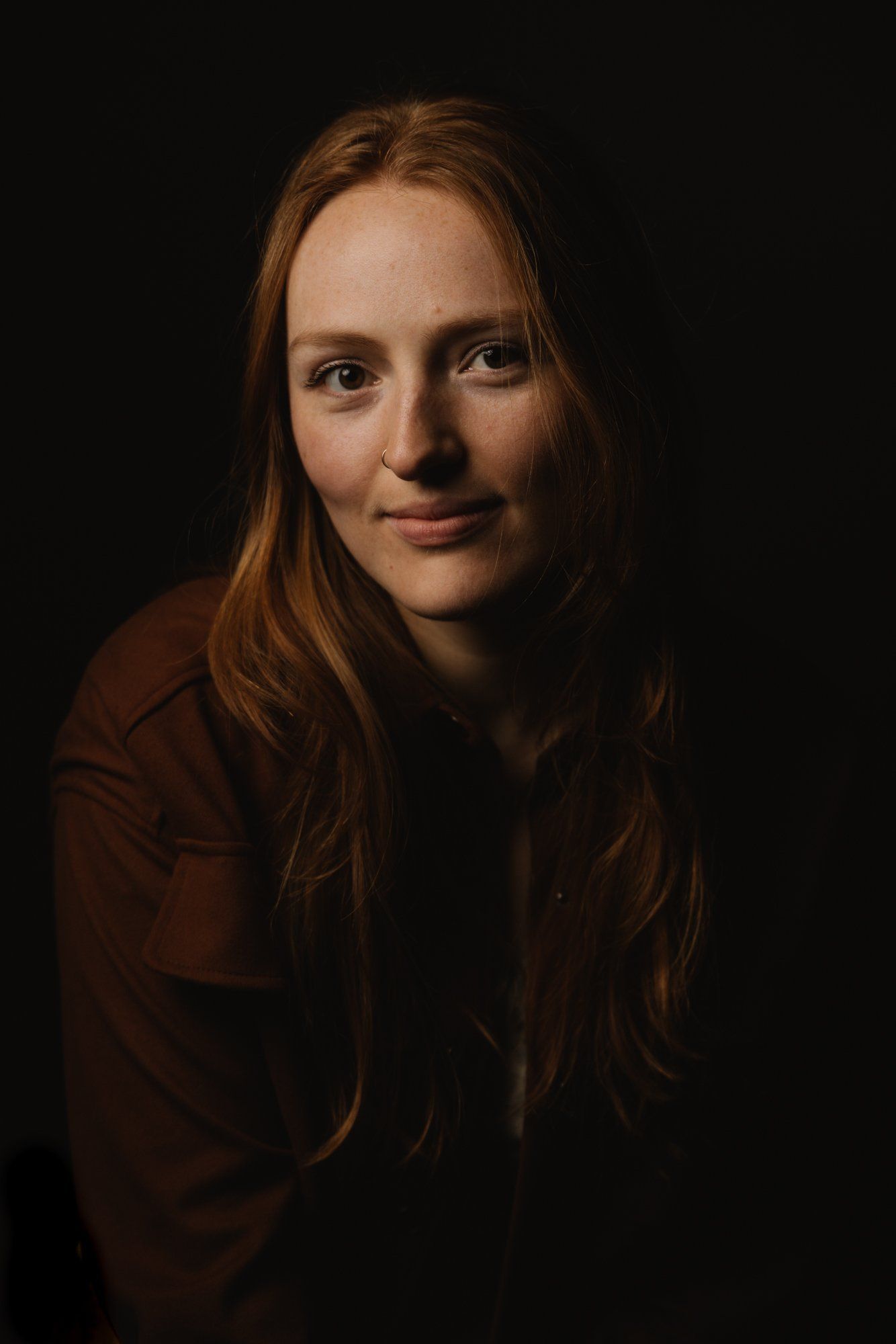 Woman with long reddish-brown hair in burgundy jacket, warmly lit against dark background, gentle smile