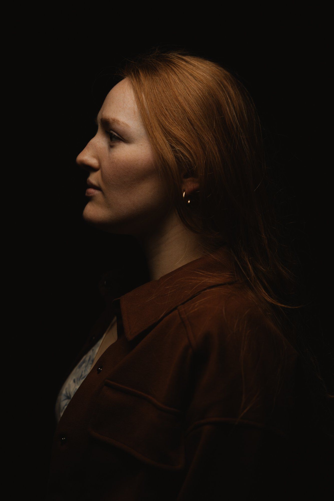 Woman in profile against black background, warm sidelight illuminating reddish-brown hair and burgundy jacket with soft, contemplative expression