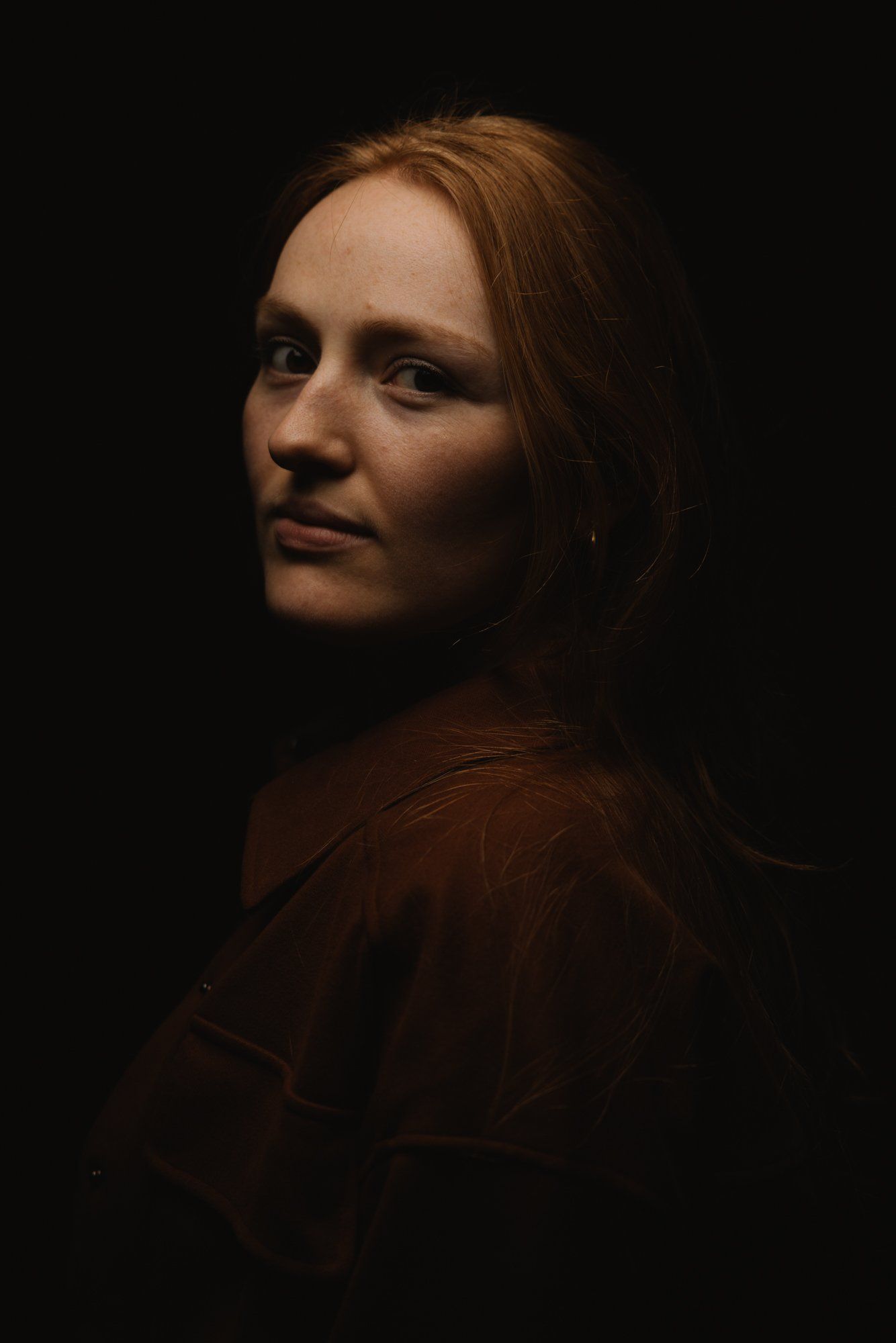 Woman with reddish-brown hair gazes over her shoulder against a dark background, dramatically lit from the side in warm golden tones