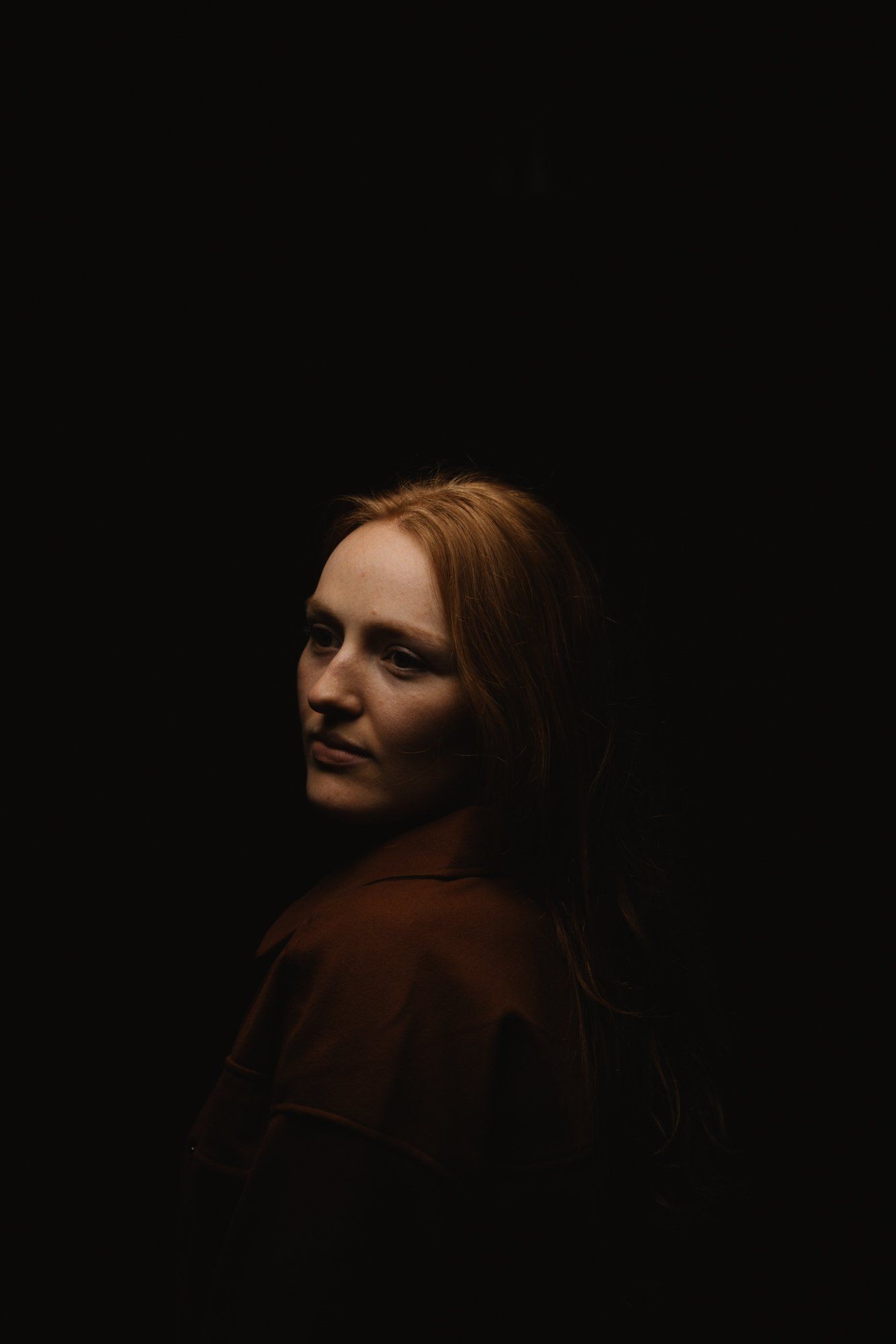 Woman with reddish-brown hair gazes over shoulder against dark background, warm side lighting sculpting her face and shoulders