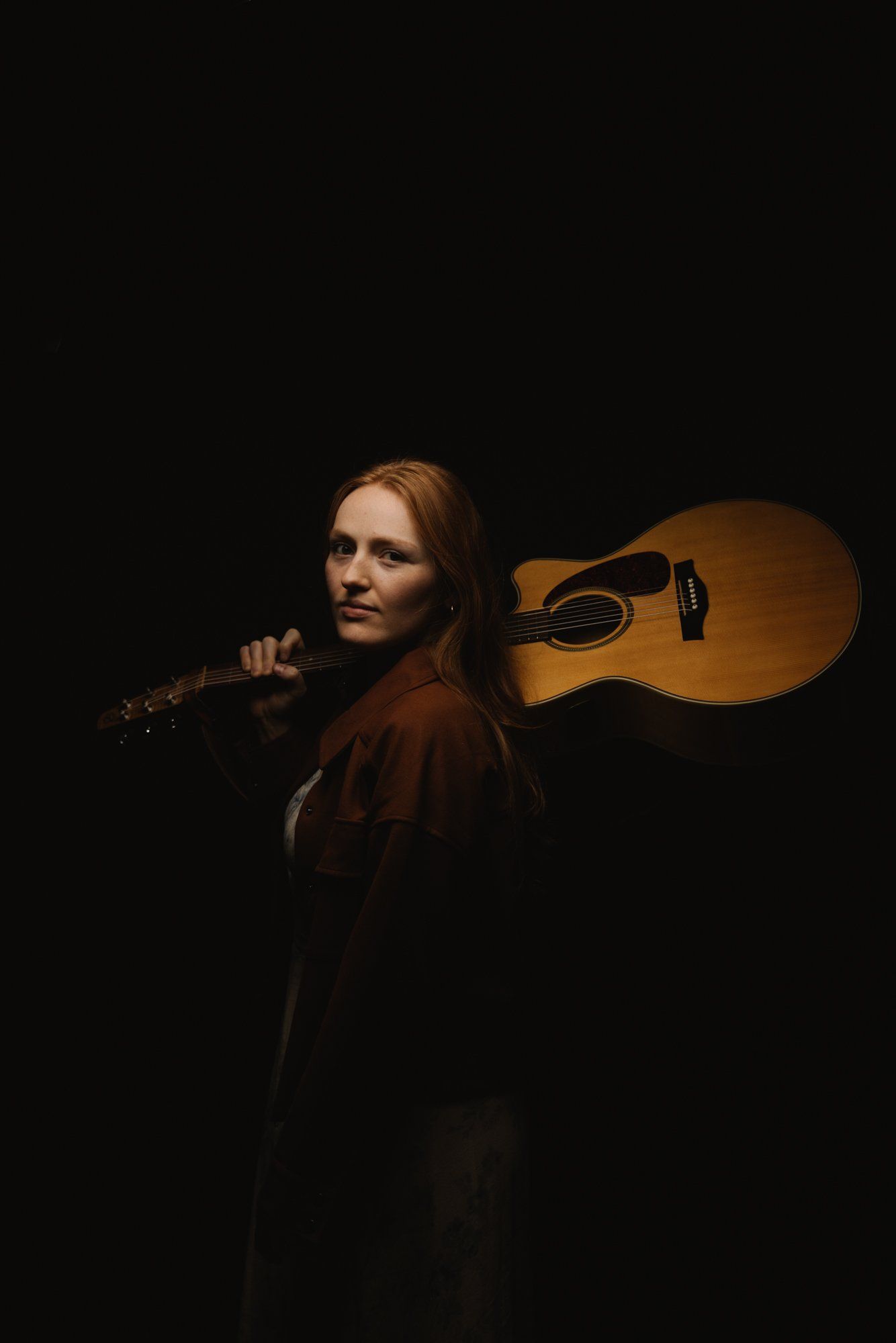 Musician with reddish hair holds an acoustic guitar against their shoulder, gazing over it in dramatic studio lighting against black background