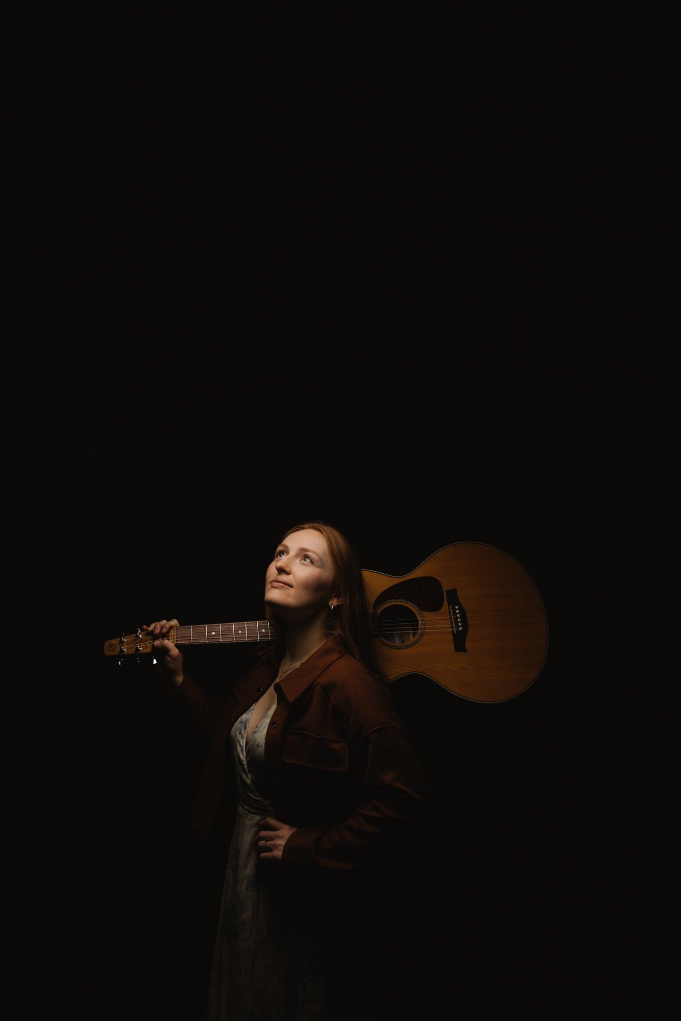 Woman in burgundy jacket holds acoustic guitar against black background, gazing upward in dramatic studio lighting