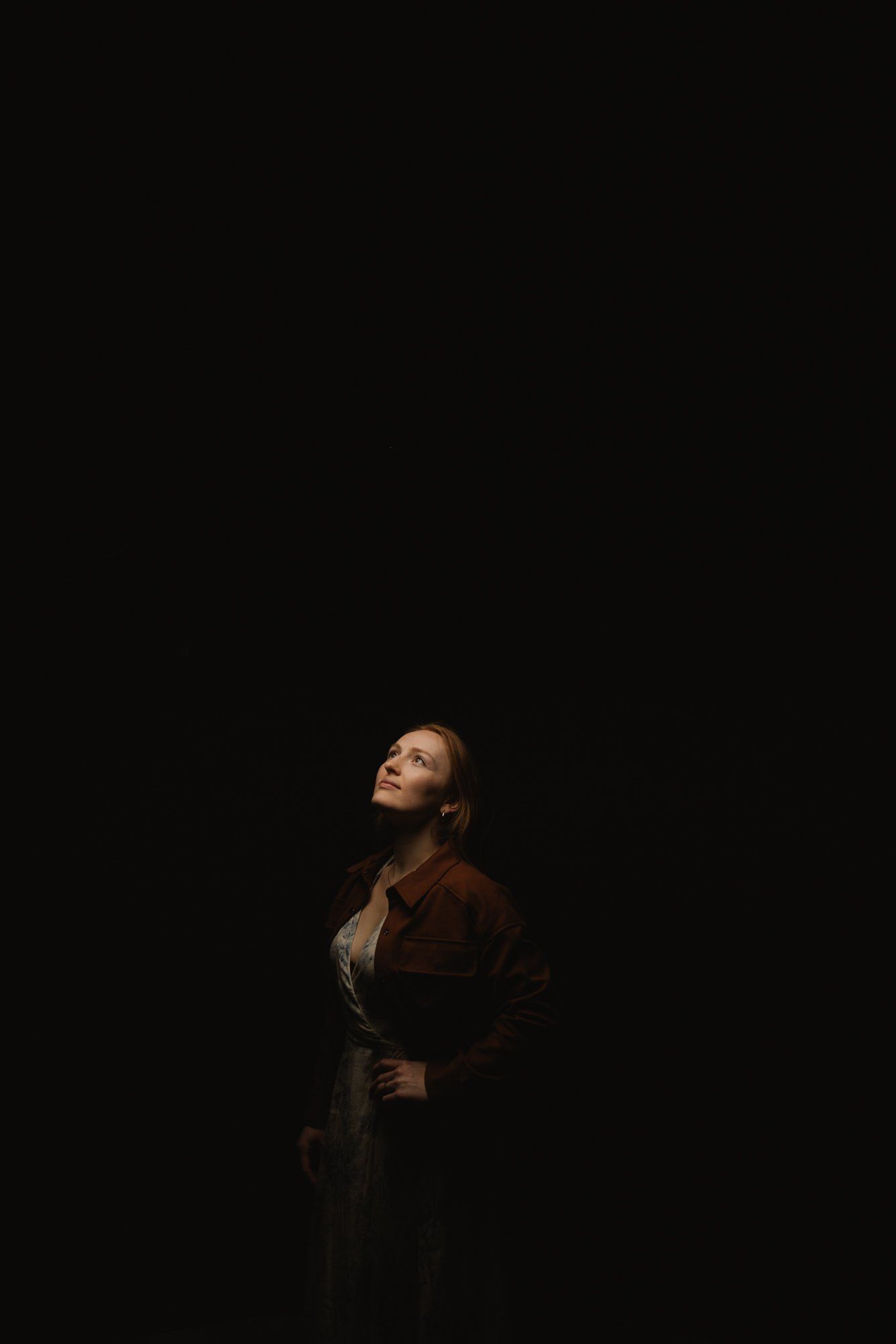 Woman in burgundy jacket gazes upward against dark background, warm side lighting sculpting her profile