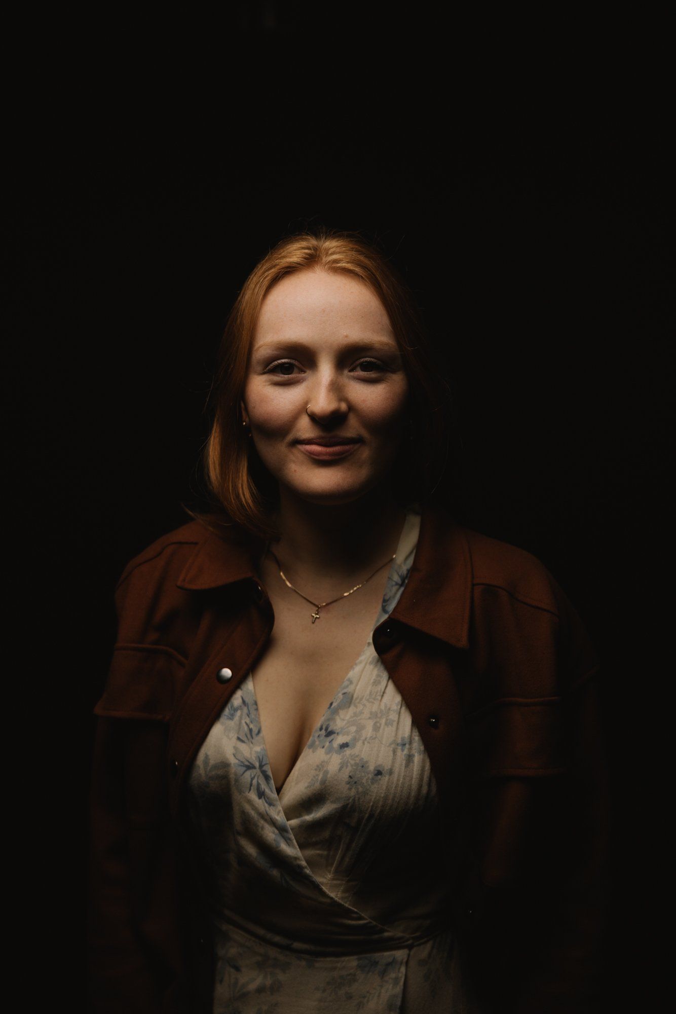 Woman with reddish-brown hair smiling softly against black background, wearing brown jacket over cream blouse with delicate necklace