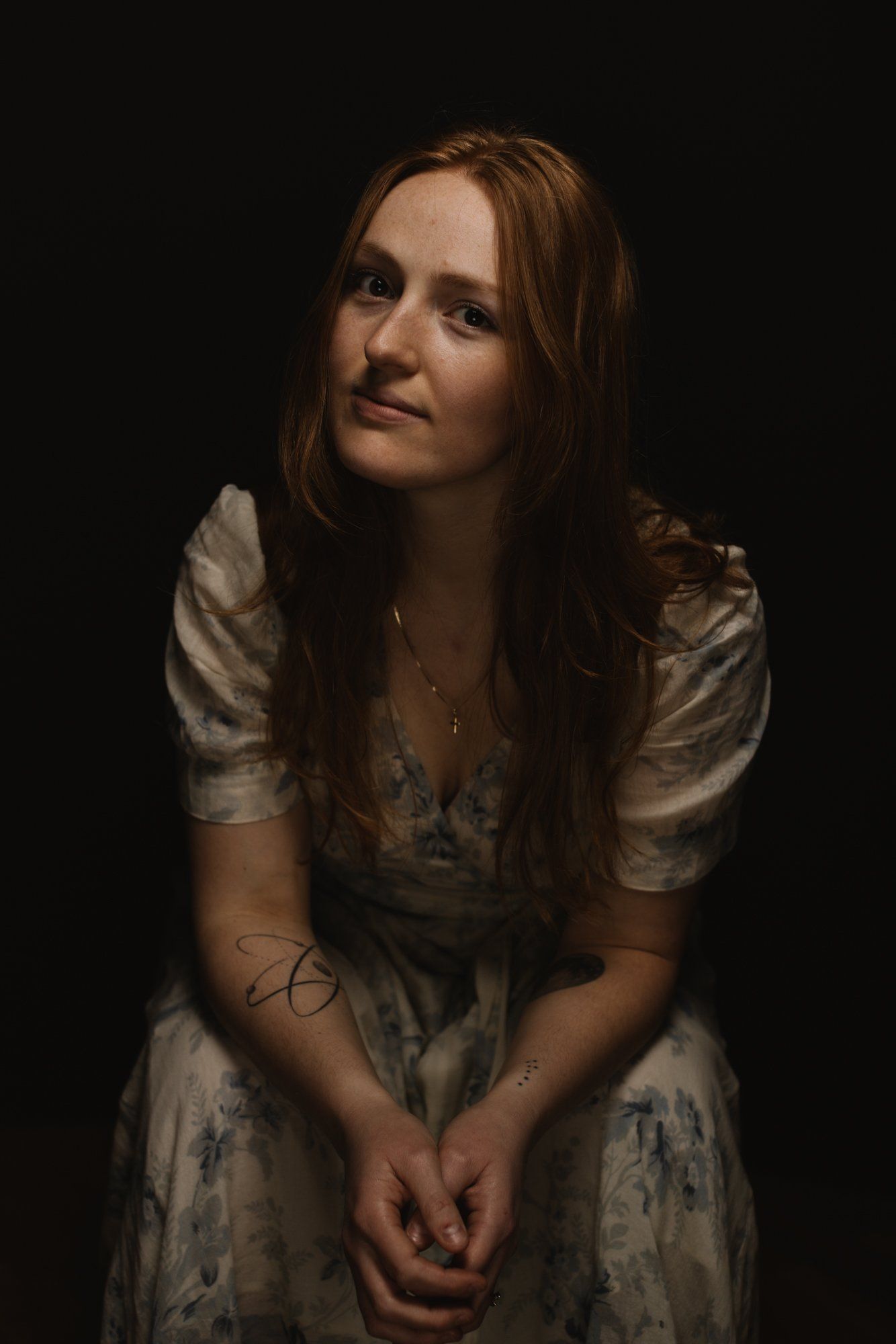 Woman with long brown hair in pale embroidered dress against dark background, soft side lighting creating contemplative mood