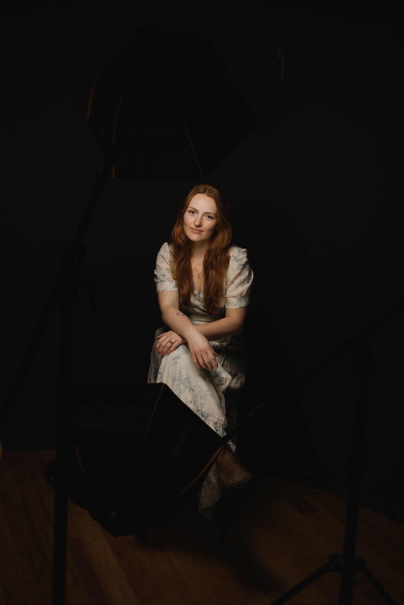 Woman in cream linen dress seated against dark background, warm light illuminating her face and contemplative expression