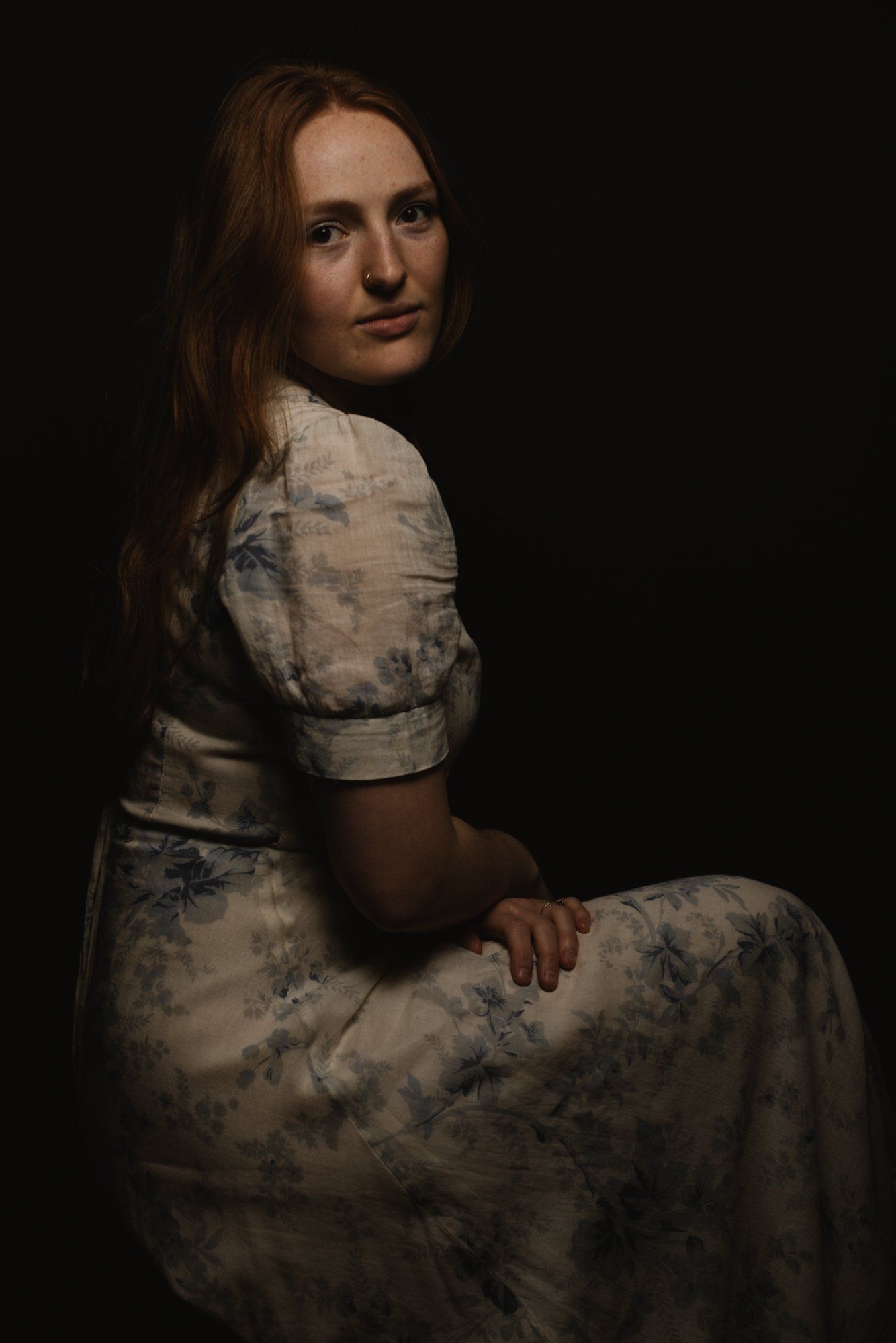 Woman in floral dress seated against dark background, gazing over shoulder with soft studio lighting