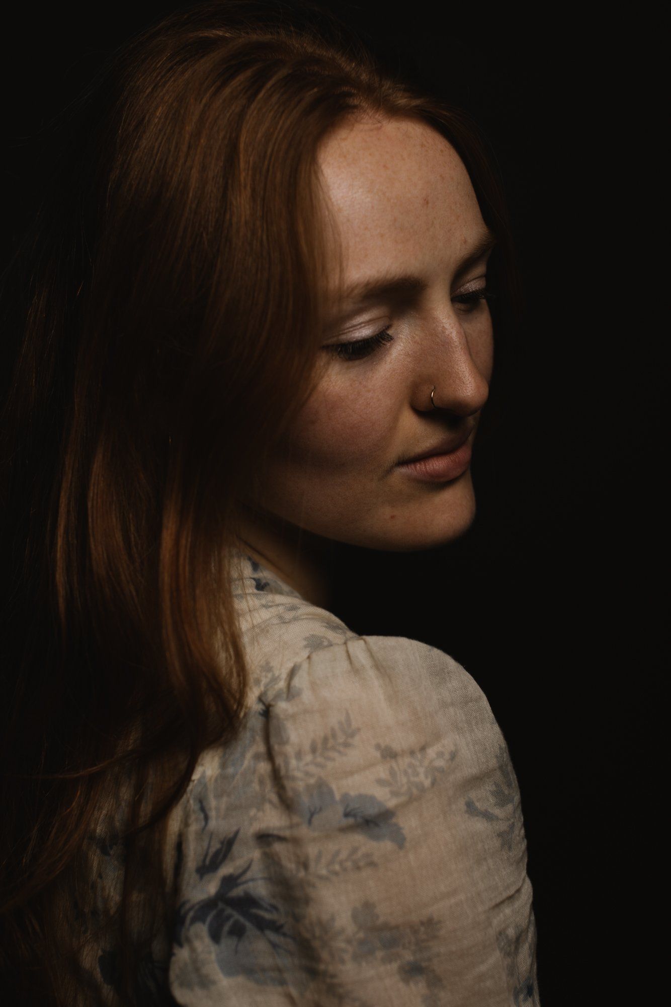 Woman in profile against dark background, soft directional light illuminating her face and patterned shirt, serene expression with downward gaze