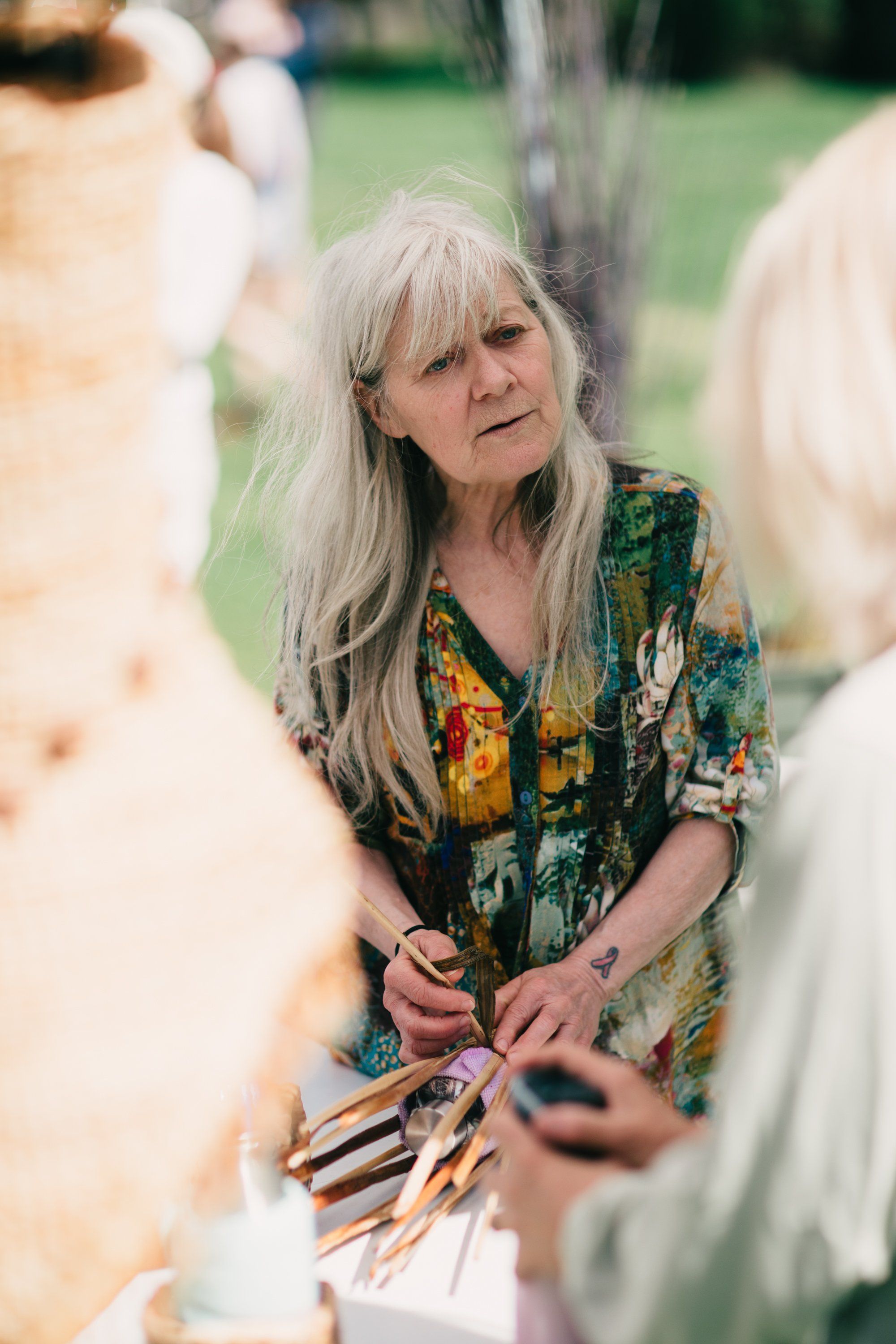 Woman with long gray hair in floral green shirt holds gardening tools while conversing with guests at a sunny farm gathering