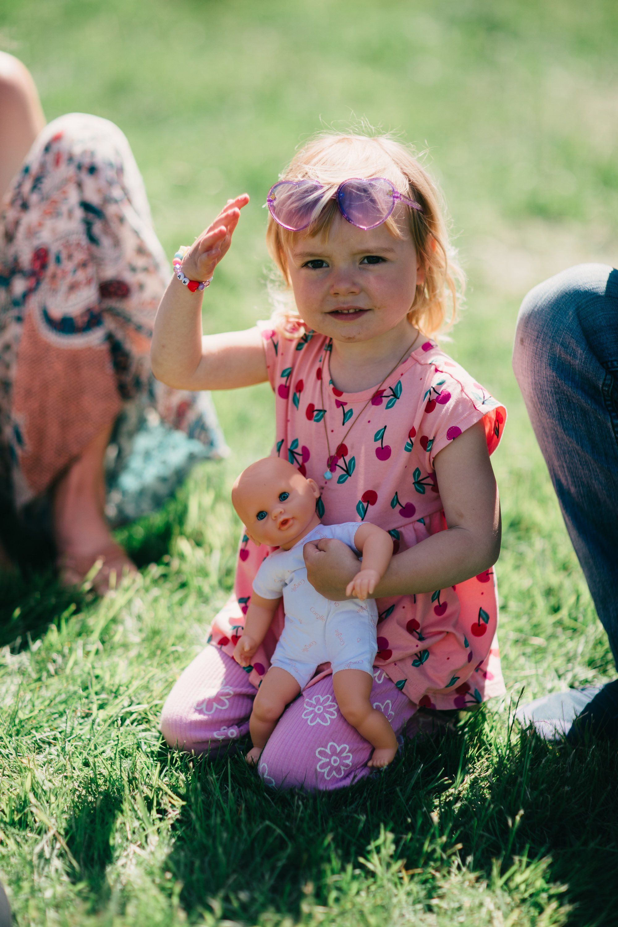 Toddler in pink floral dress holding a doll sits on grass surrounded by adults, wearing oversized sunglasses and smiling at camera