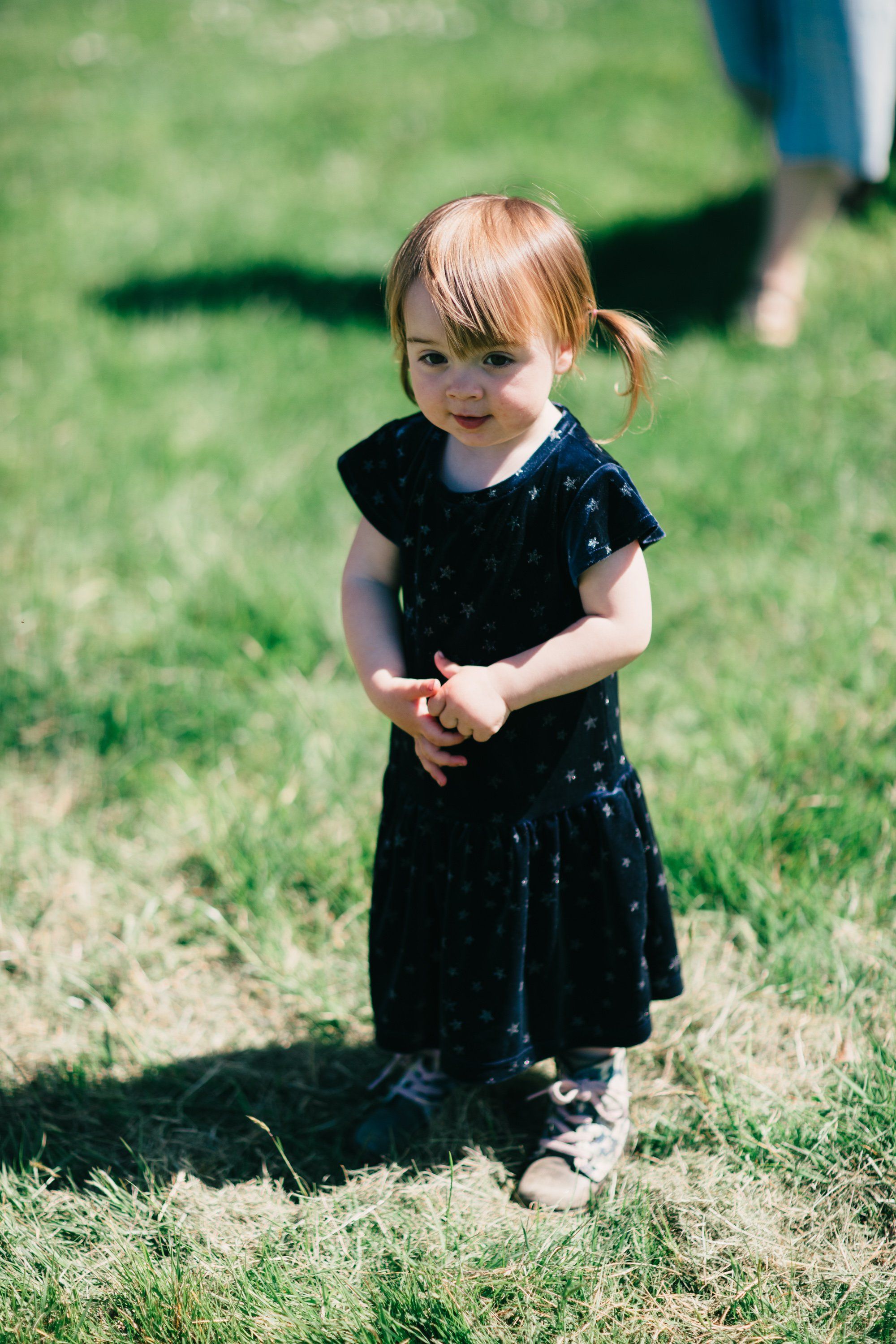 Toddler in black polka-dot dress stands on sun-dappled grass, looking down thoughtfully at her clasped hands