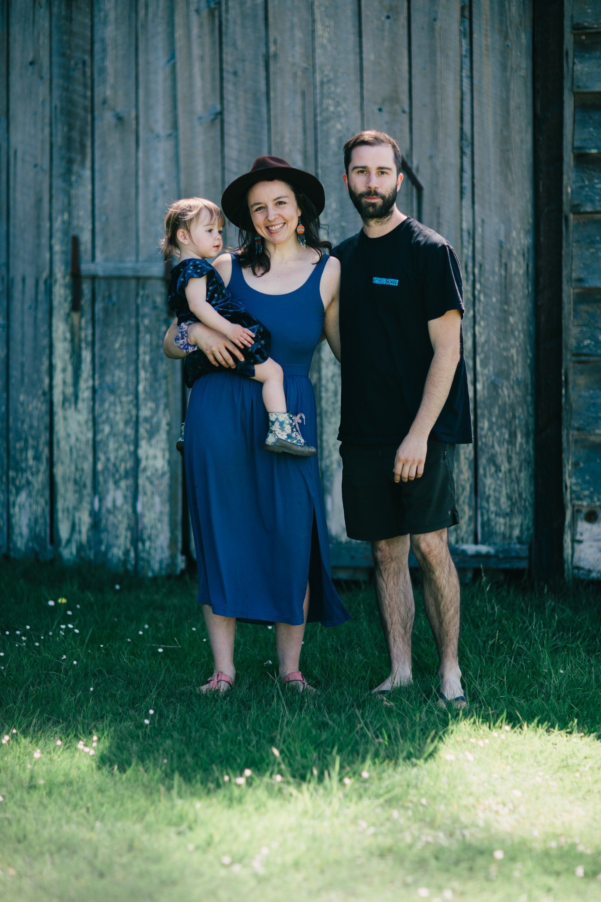 Family of three standing on grass before weathered blue barn doors, soft natural light, woman in blue dress holding toddler, man in black shirt beside them