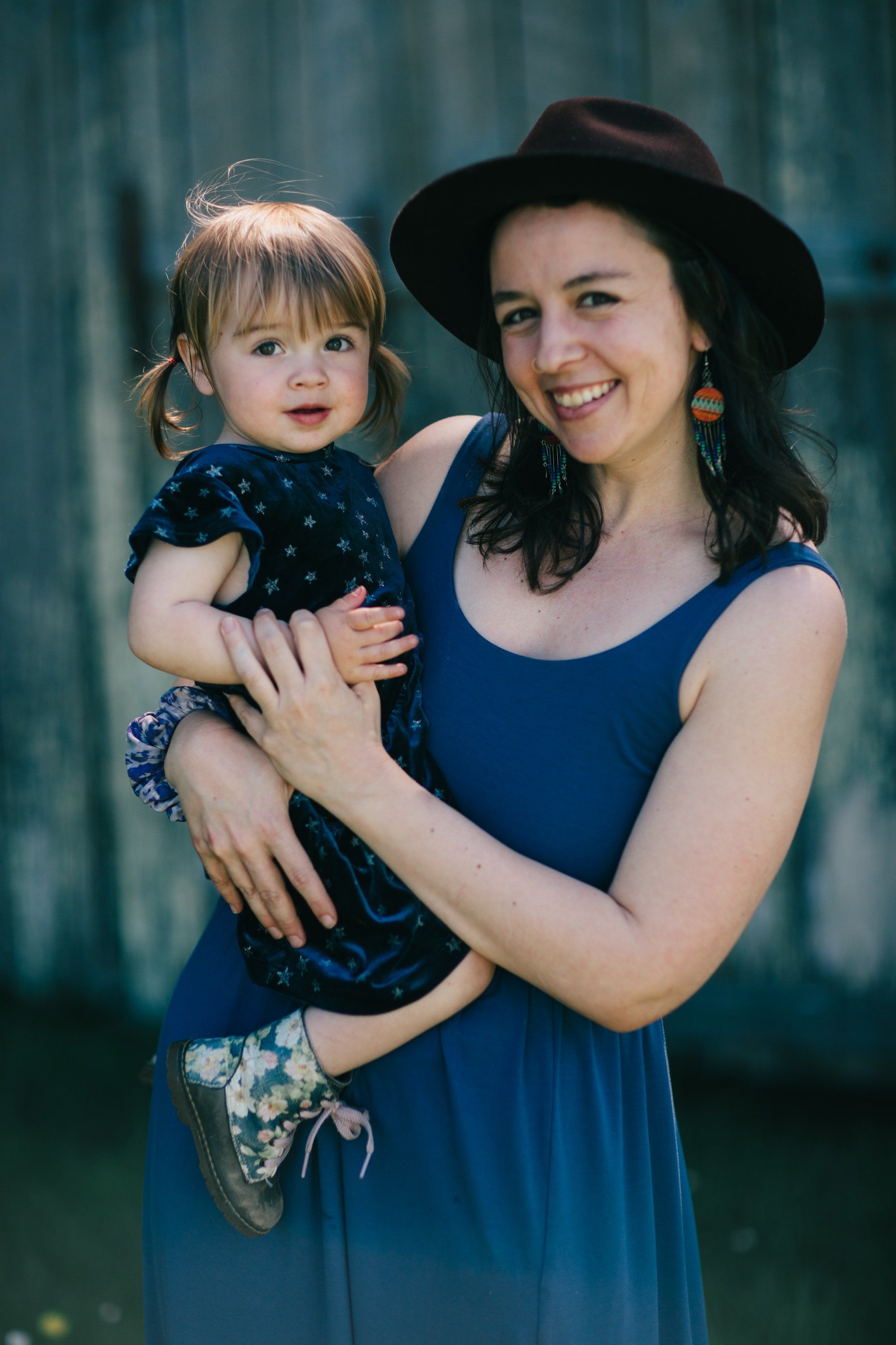 Woman in blue dress and black hat holding smiling toddler, both looking at camera with warm natural light and soft teal background