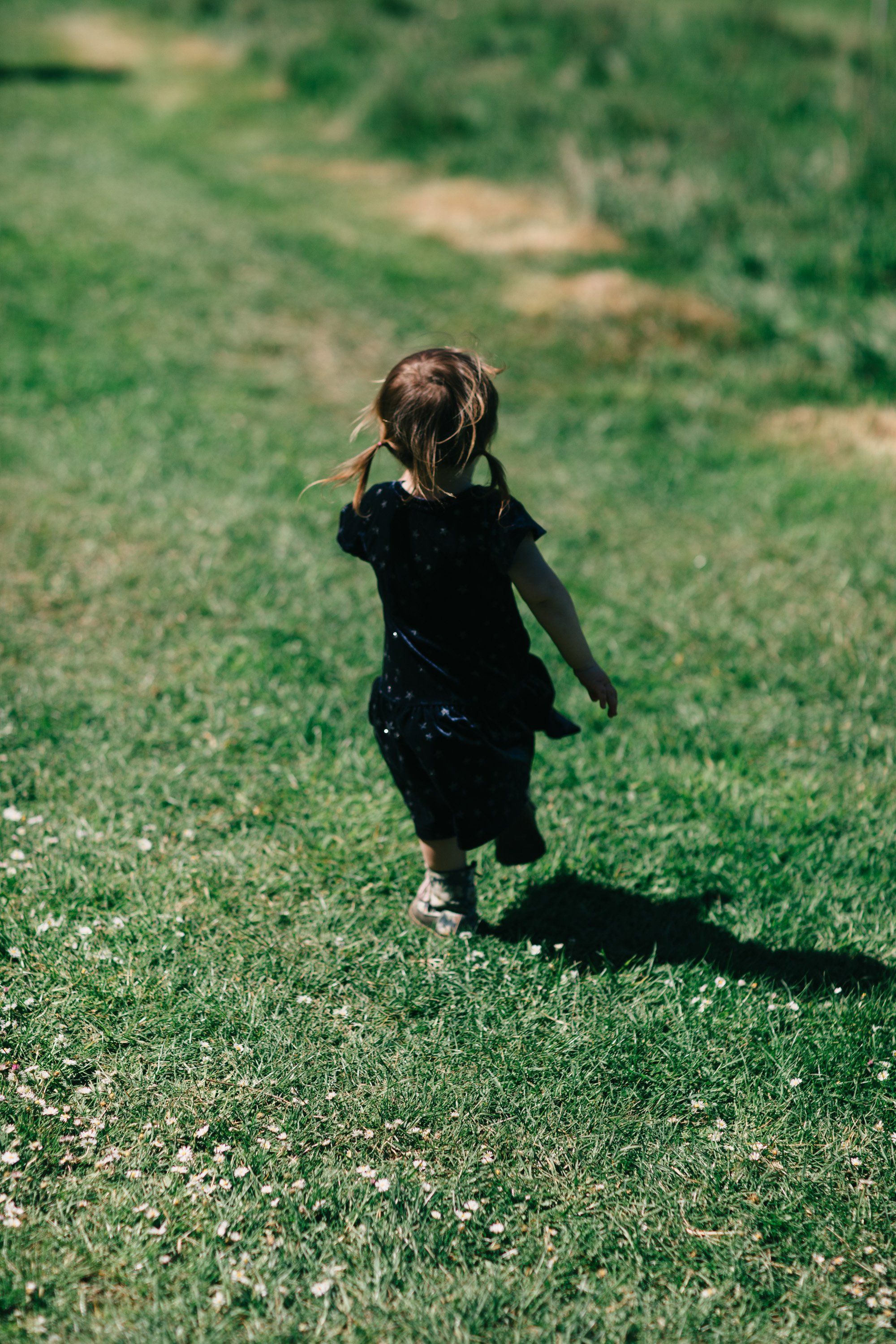 Child in dark clothing running through sunlit grass field, casting a long shadow on a bright summer day
