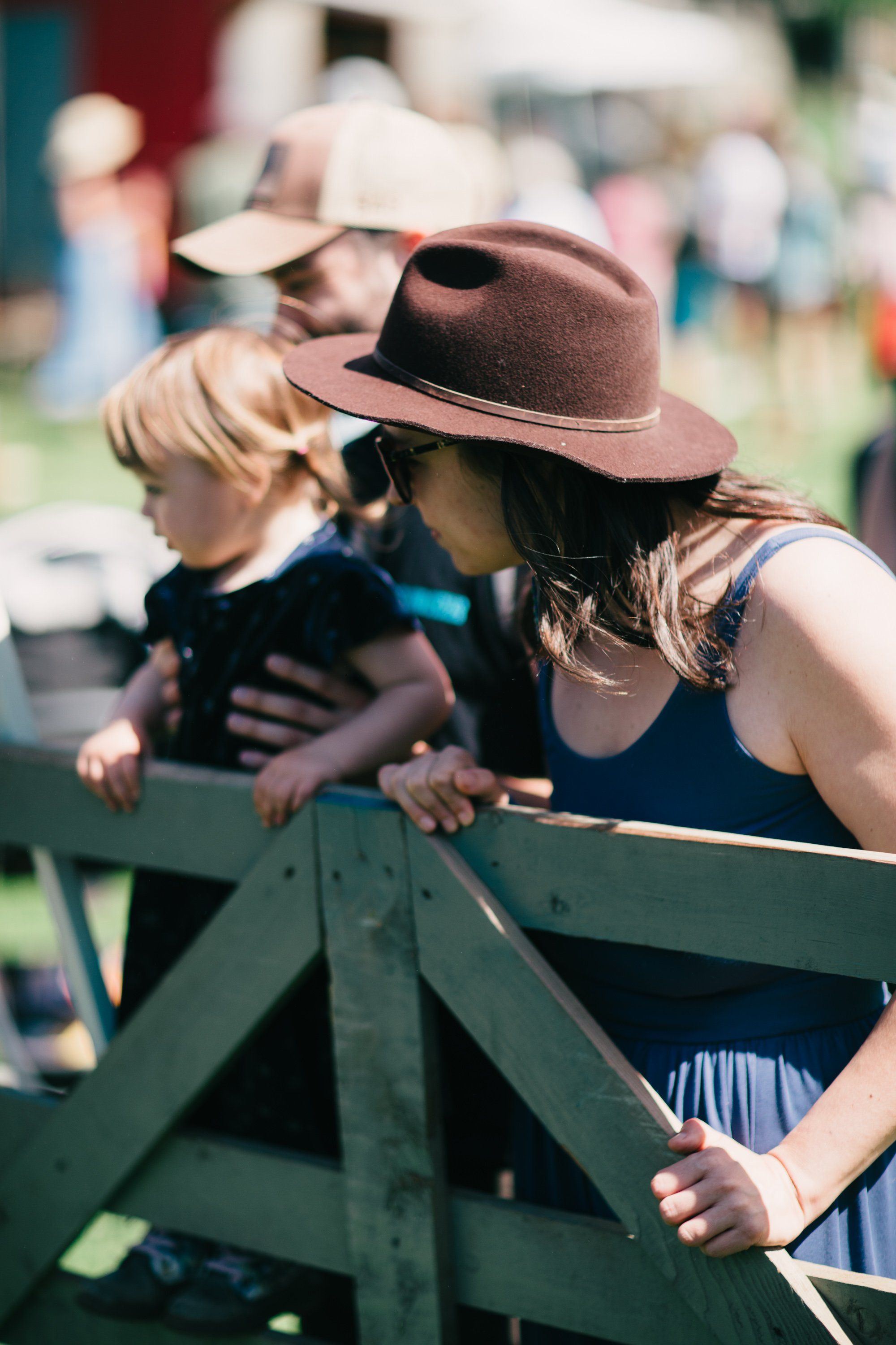 Woman in brown hat and blue dress leans over wooden fence railing with young child at sunny farm gathering
