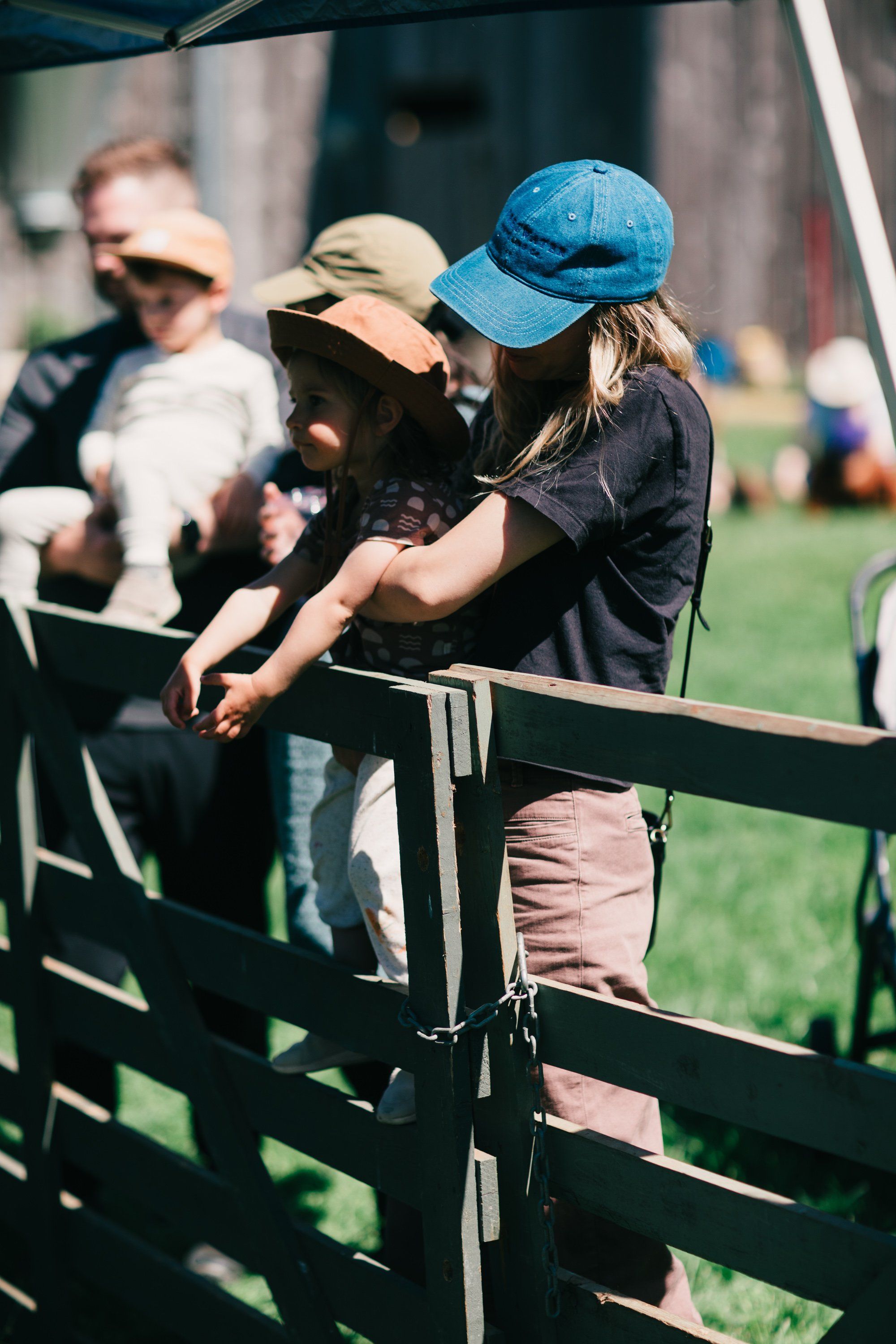 Child in blue cap leans over wooden farm fence in bright sunlight, examining something below while adults watch from the background