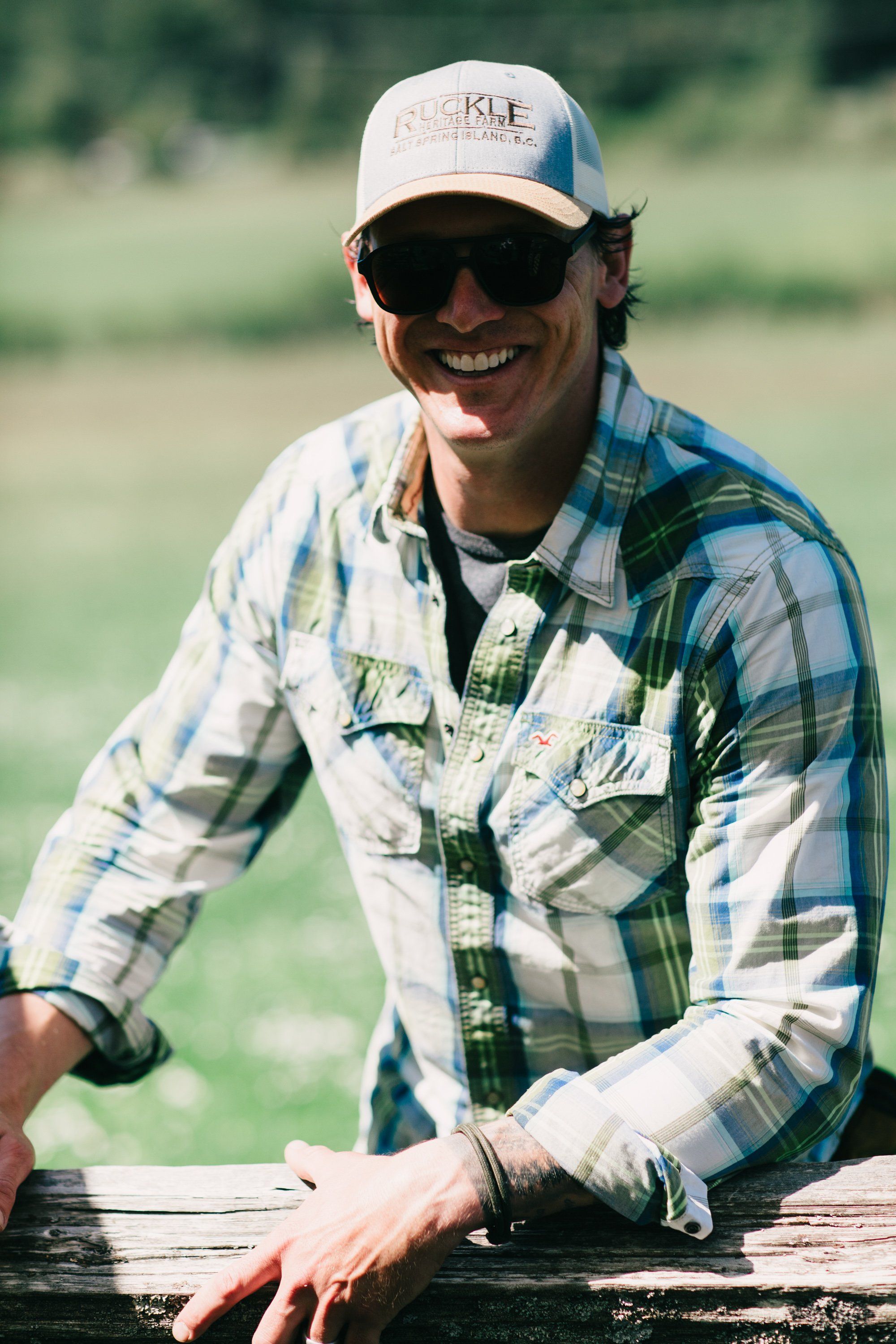 Man in blue plaid shirt and cap smiles at camera while leaning on wooden fence in sunlit pastoral field