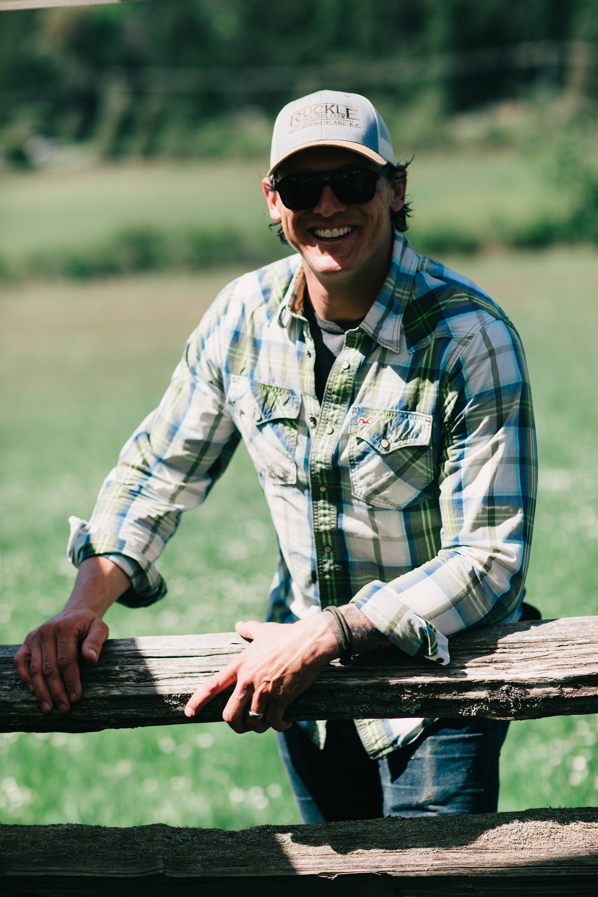 Man in plaid shirt and cap leaning on wooden fence rail with green pasture behind him in golden afternoon light
