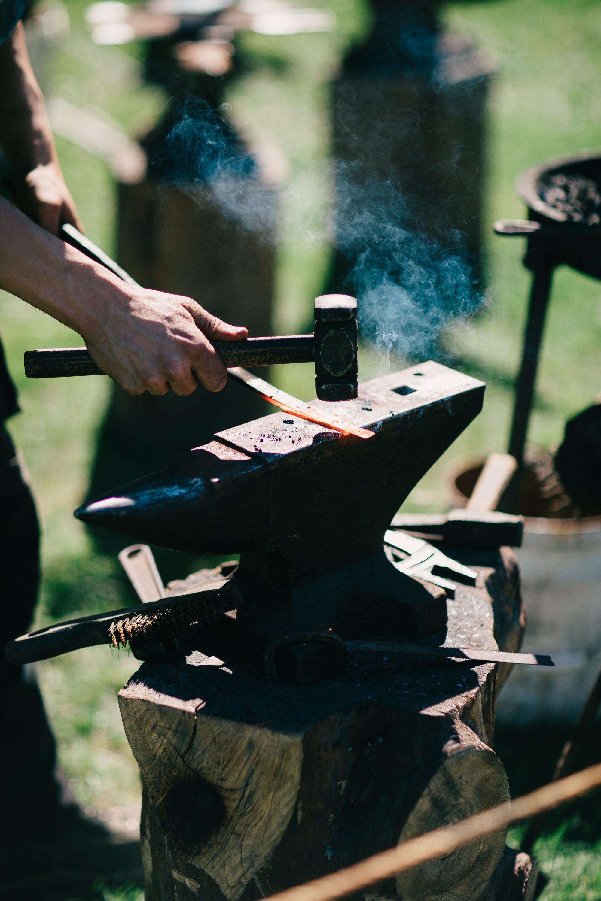 Blacksmith's hands working metal at an anvil with hammer and tongs, smoke rising in bright sunlight on a farm
