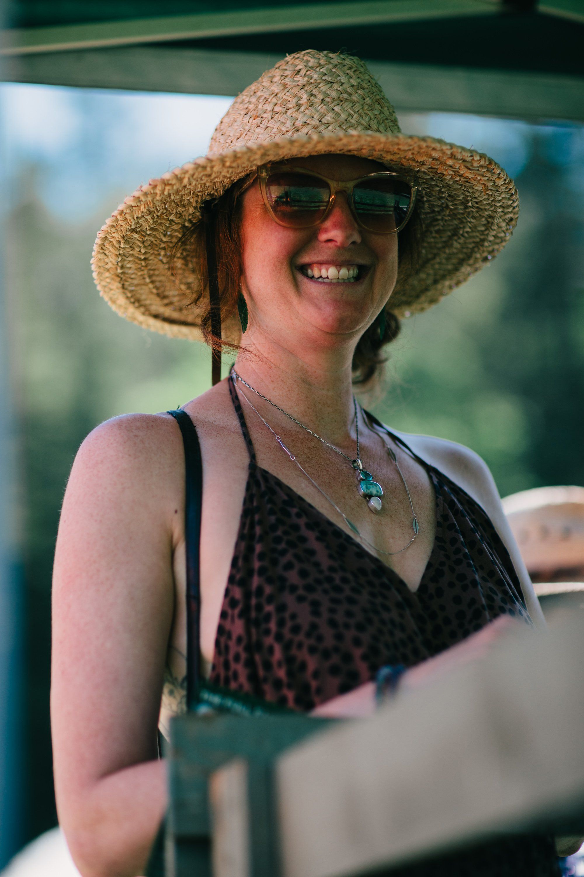 Woman in straw hat and red-tinted sunglasses smiles warmly at Ruckle Farm, golden afternoon light filtering through trees behind her
