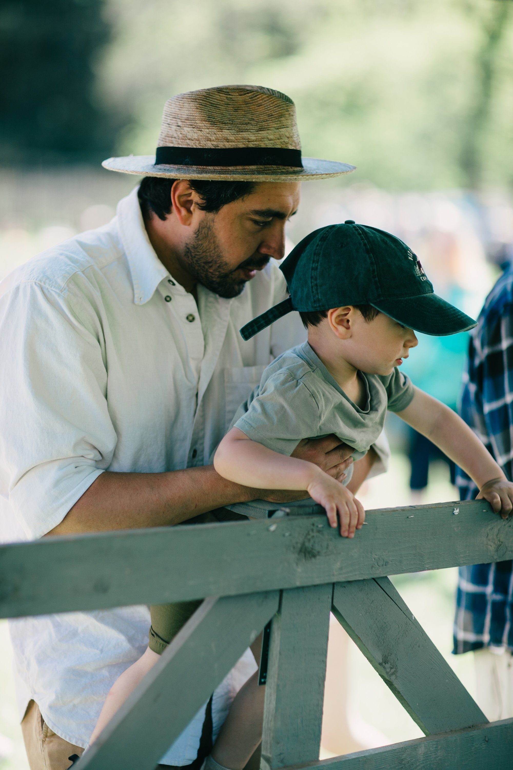 Man in straw hat and boy in cap lean together over weathered wooden fence rail in golden afternoon light