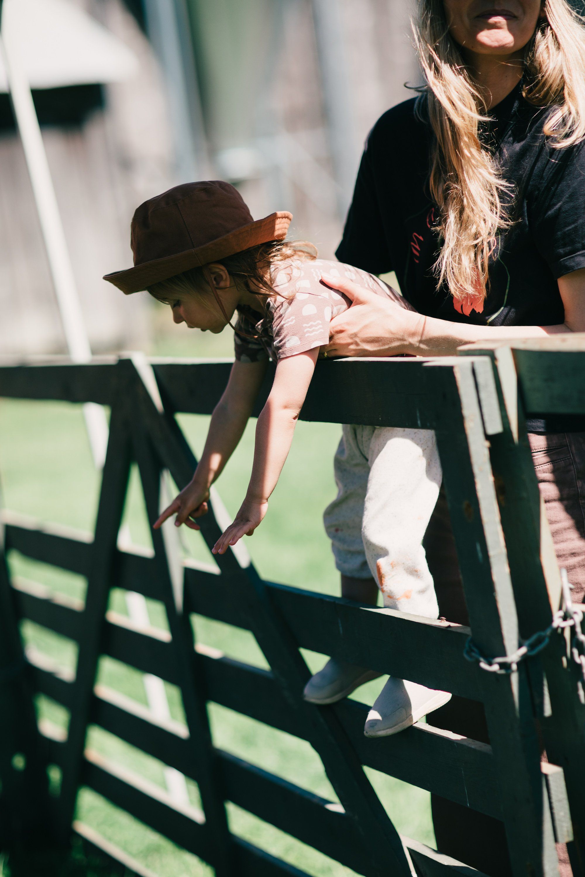 Child in a brown hat leans over a weathered fence rail in dappled sunlight while an adult watches nearby