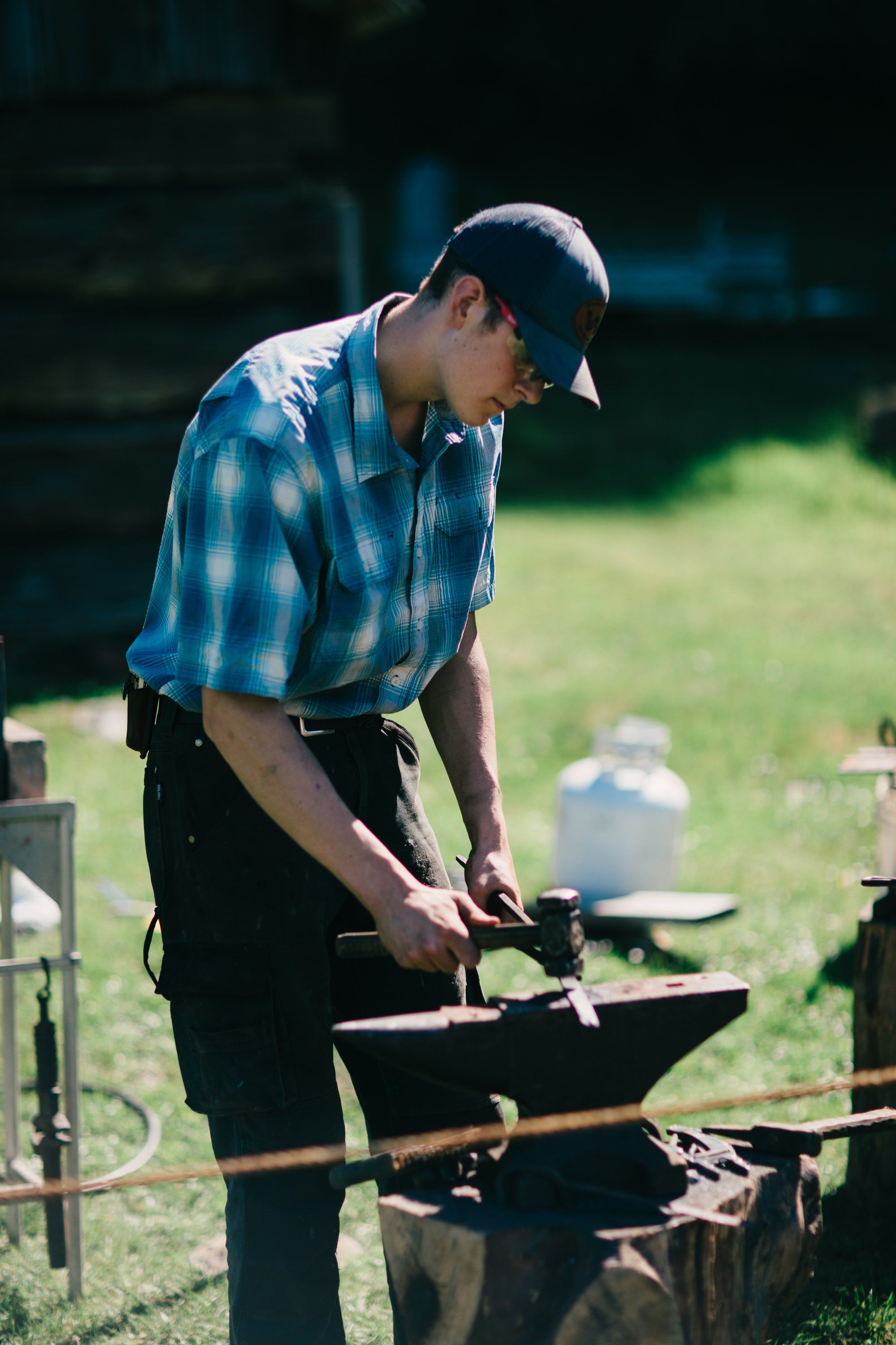 Man in blue plaid shirt and cap works at a blacksmith's anvil in bright sunlight on a grass field
