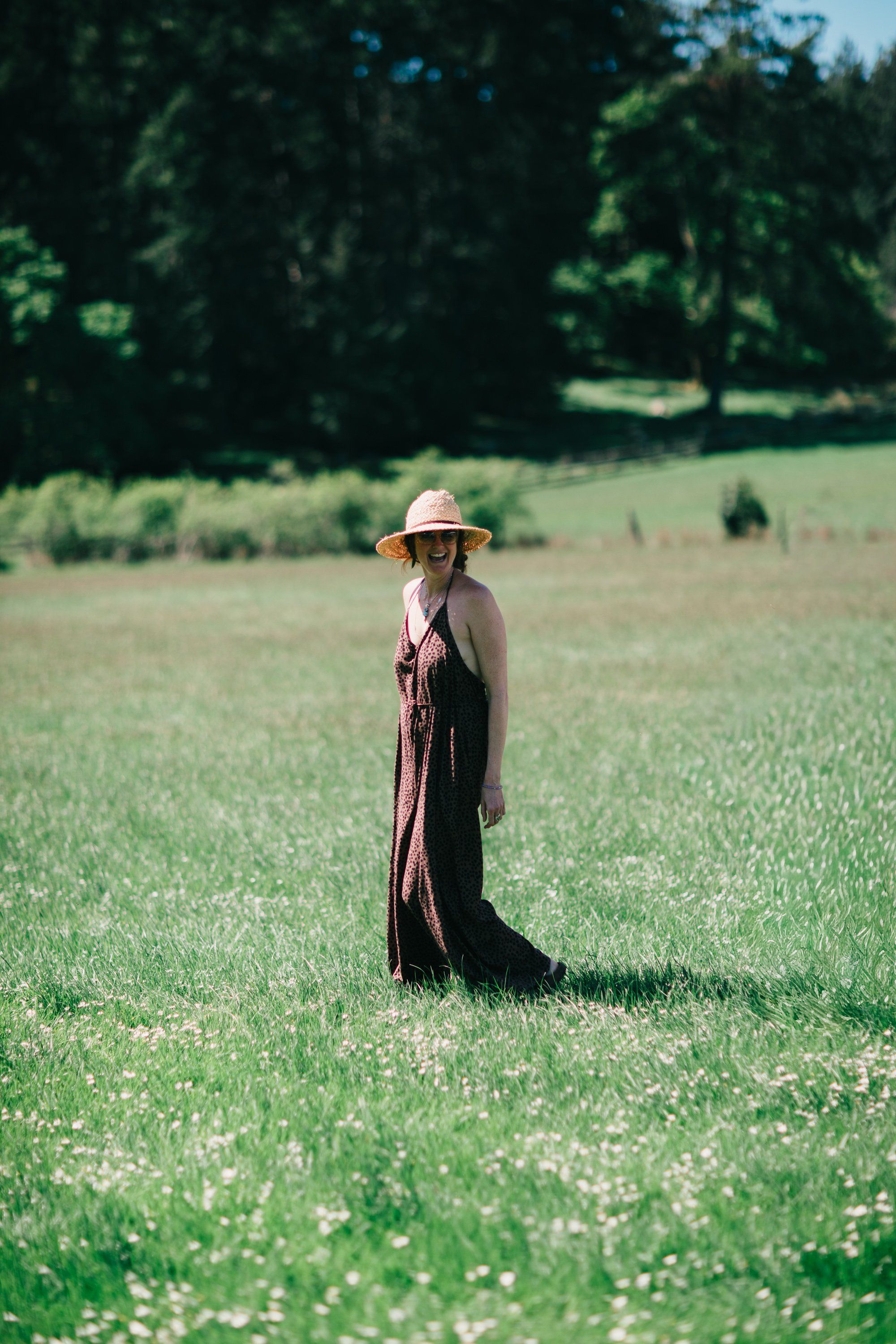 Woman in wide-brimmed hat and flowing dark dress standing alone in sunlit wildflower meadow with forested hills beyond