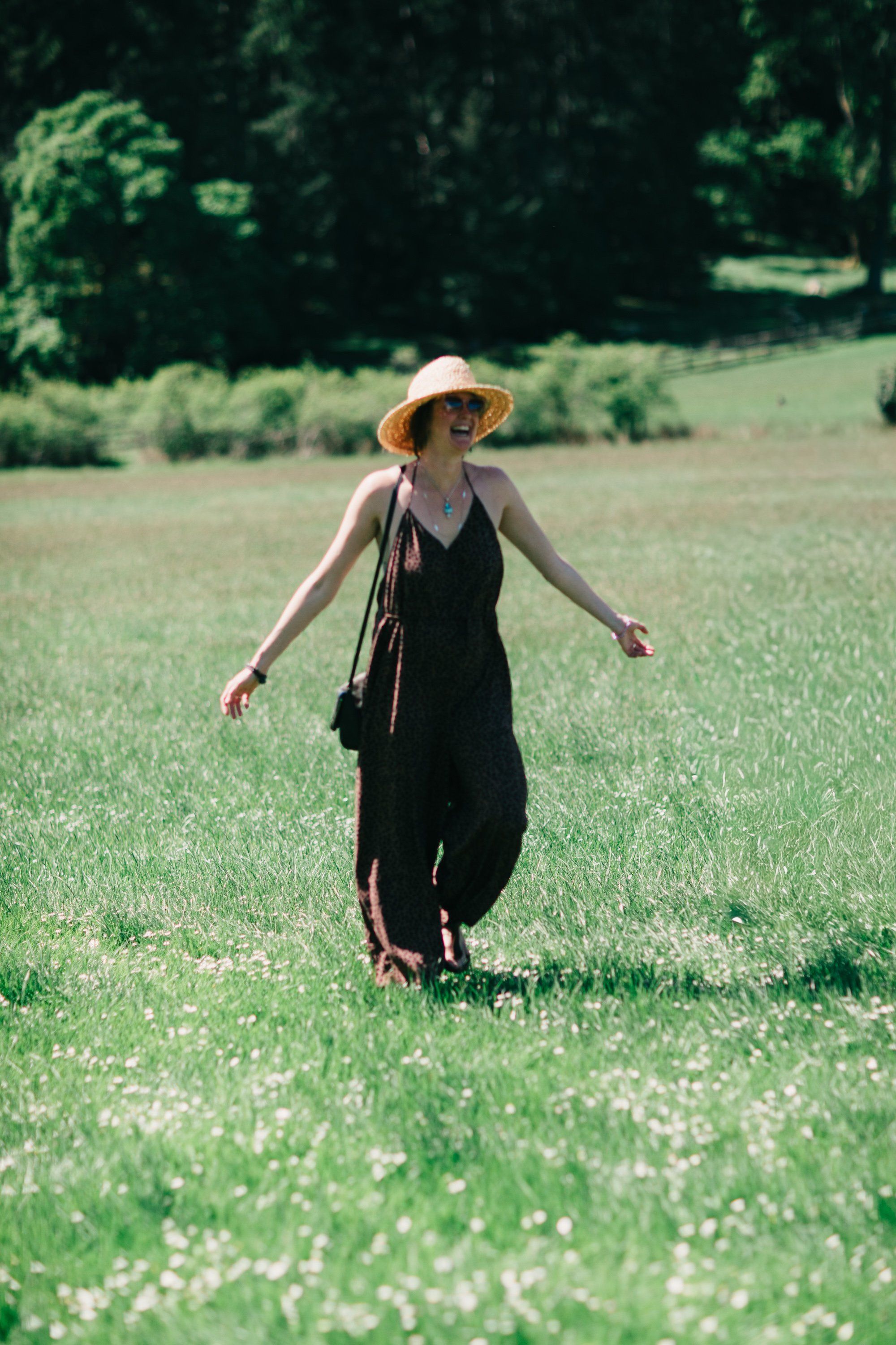 Woman in black dress and straw hat walking through a wildflower meadow with forested hills beyond