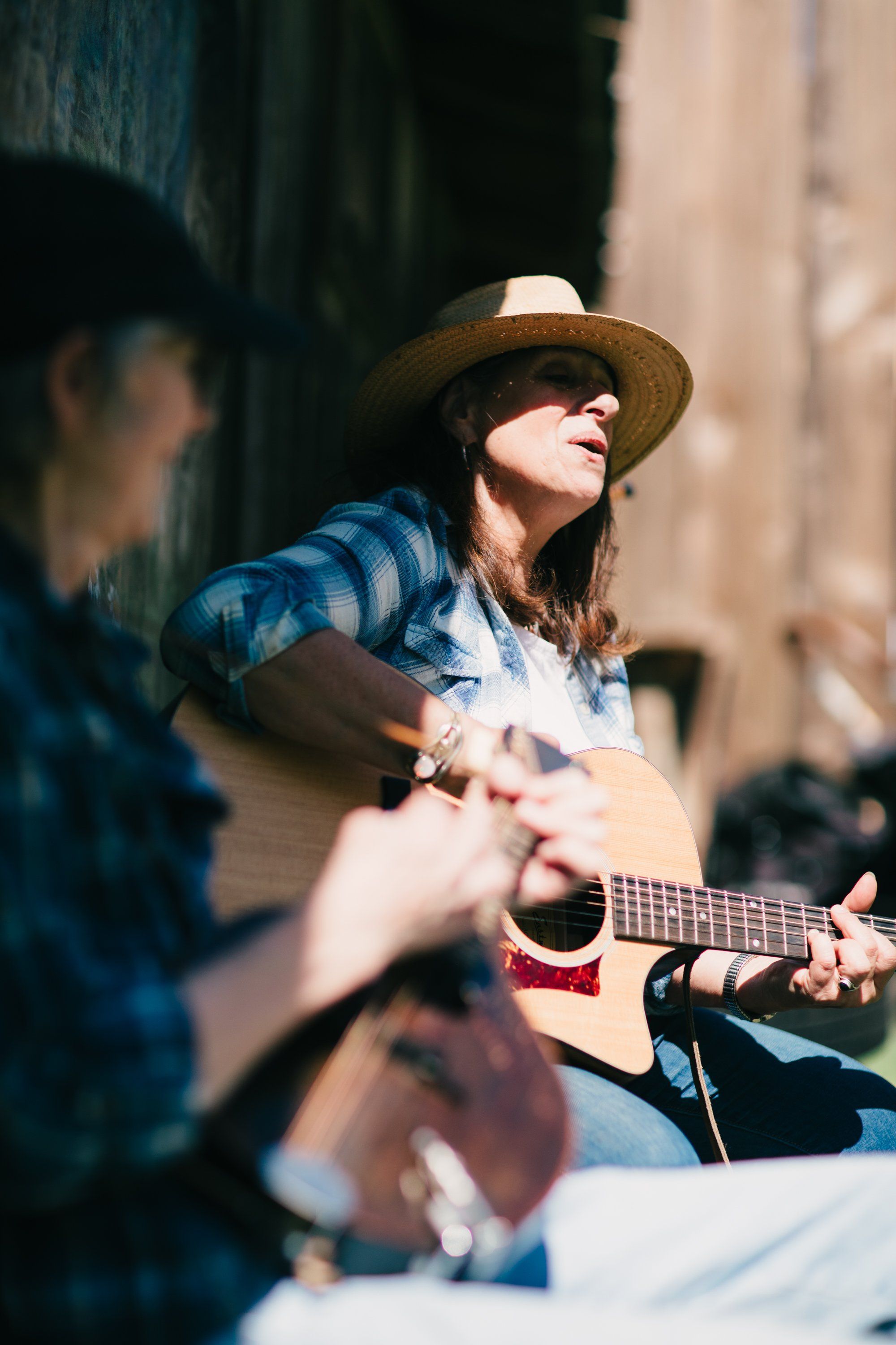 Musician in straw hat playing acoustic guitar beside rustic barn wall, dappled sunlight casting shadows across face and instrument