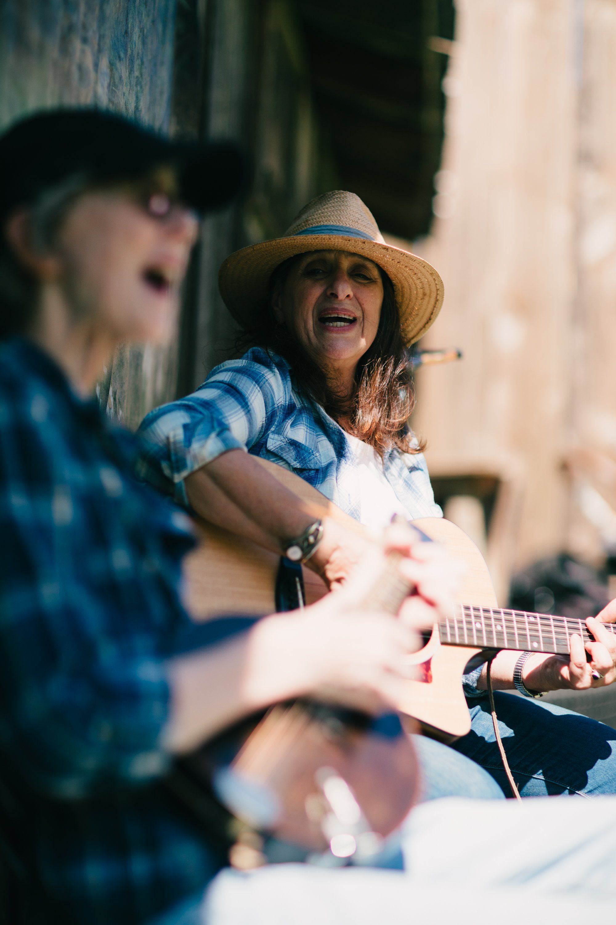 Woman in straw hat and blue plaid shirt plays guitar outdoors, smiling while seated beside a man with blurred focus in warm sunlight