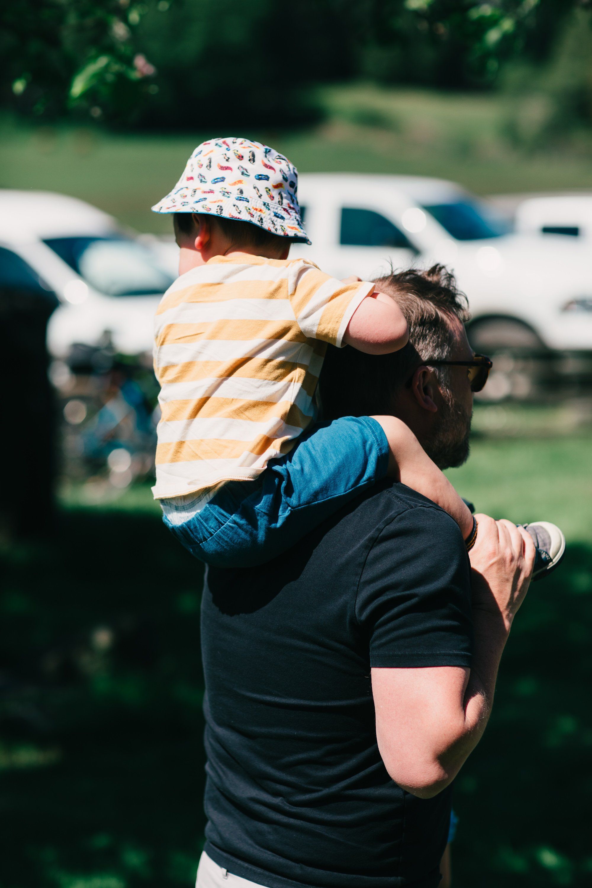 Adult holding young child on shoulders in yellow striped shirt and patterned hat at sunny farm with parked vehicles and green fields