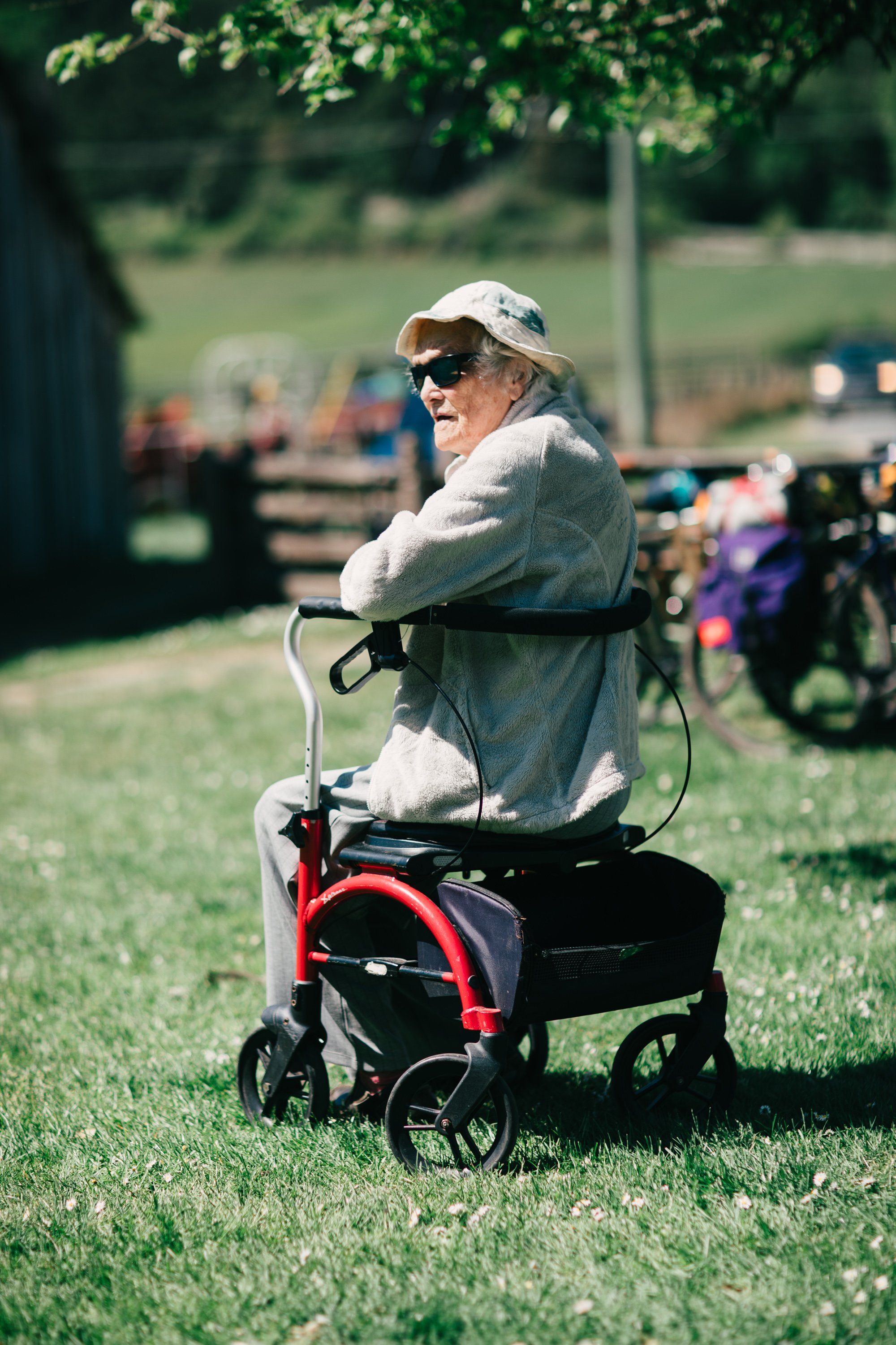 Elderly woman in sunglasses and cap pushes a red wagon across sunny farmland grass, watching the action at a distant fair