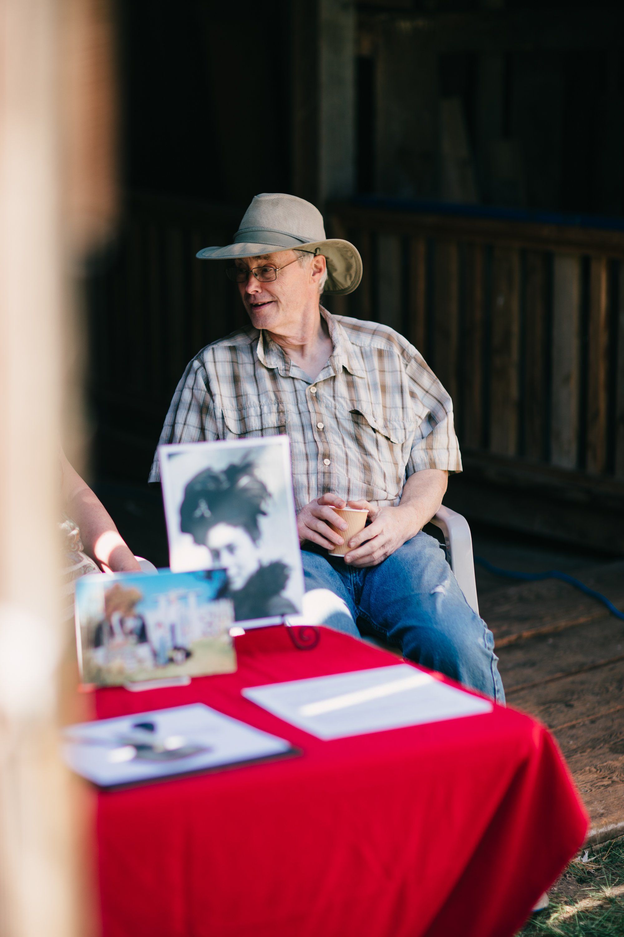 Man in cowboy hat and plaid shirt sits at red-draped table with farm photographs, golden afternoon light streaming across wooden barn interior