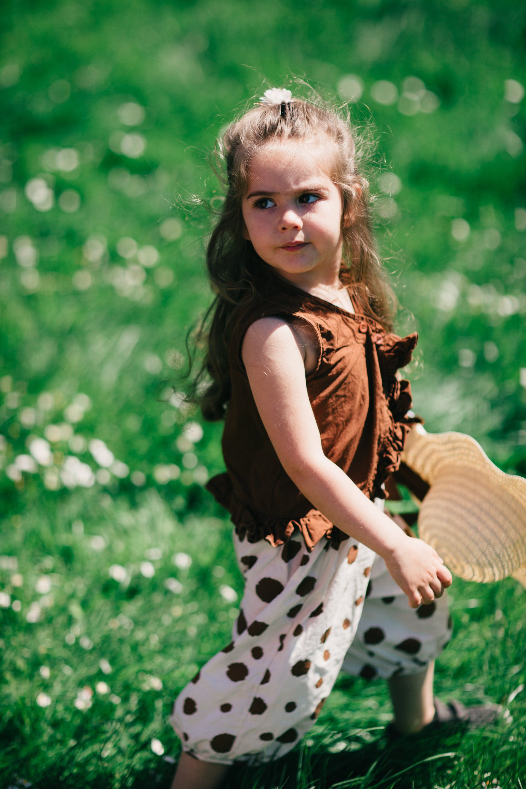 Young child in brown vest and polka-dot leggings standing in sunlit meadow with soft green bokeh background