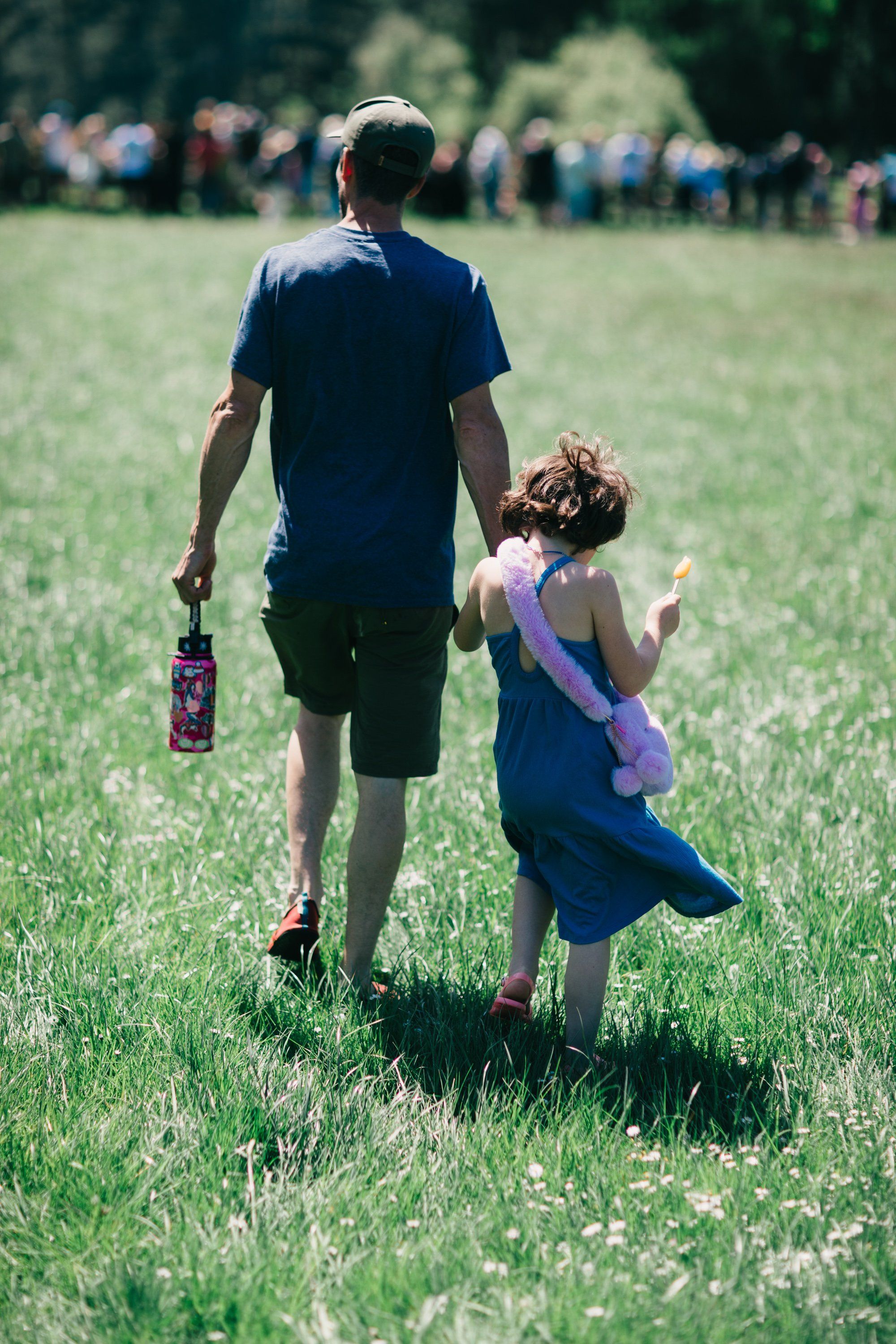 Adult and child walking hand-in-hand through a sunny meadow with a crowd gathered in the distance