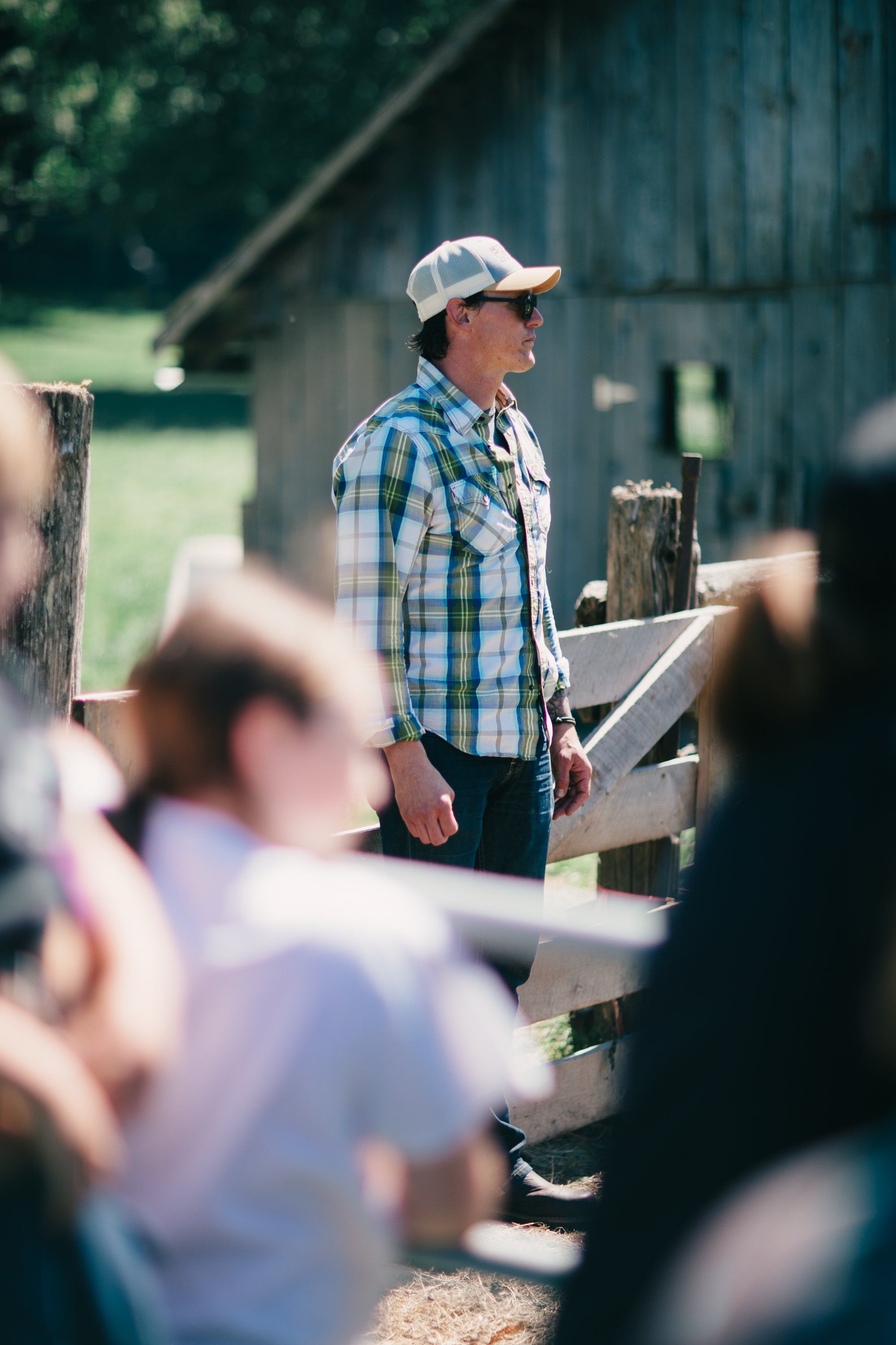 Farmer in blue plaid shirt and white cap speaks to gathered visitors at wooden farm fence in bright sunlight