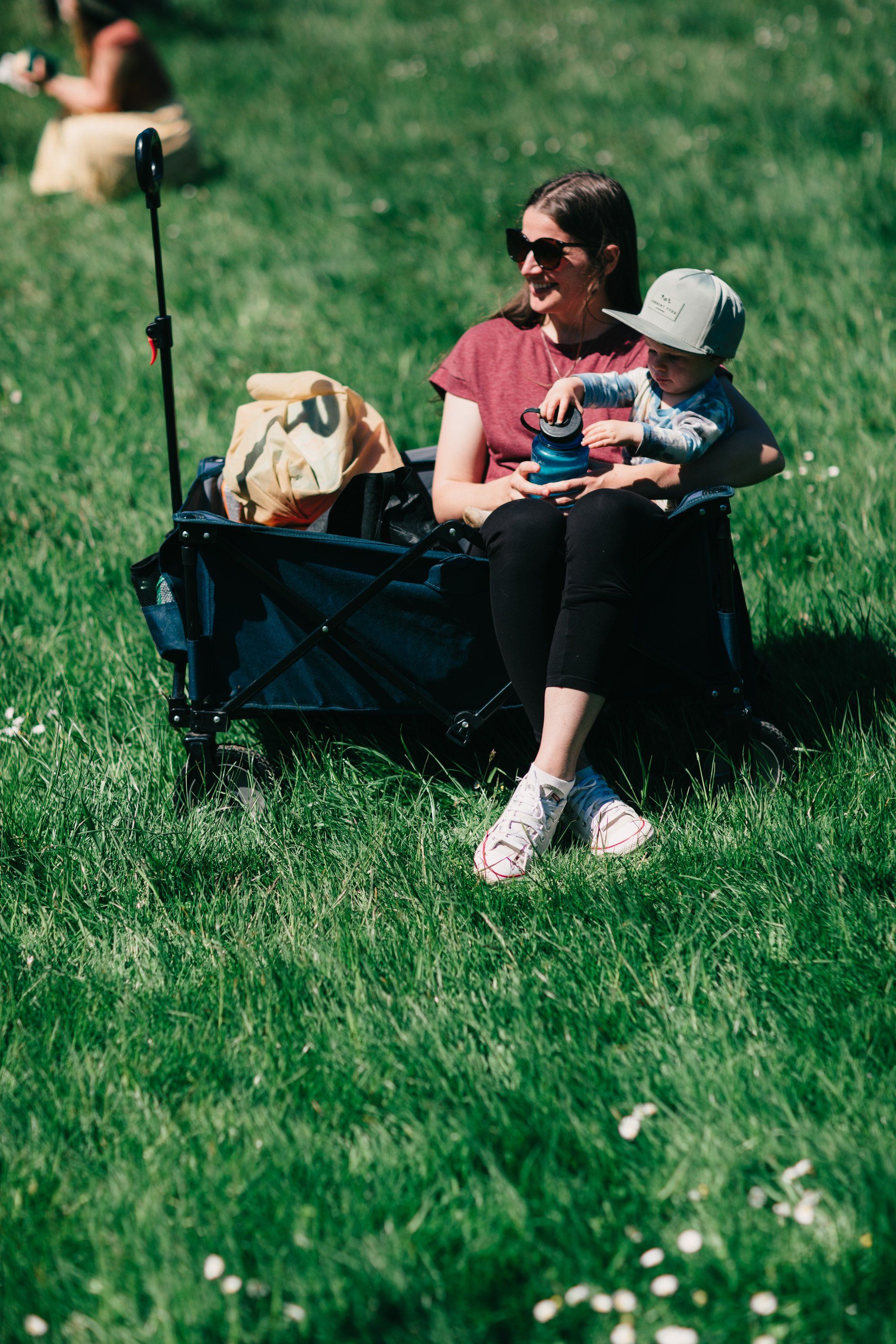 Woman in sunglasses sitting in blue wagon on grass with two young children, one in her lap, gentle afternoon light